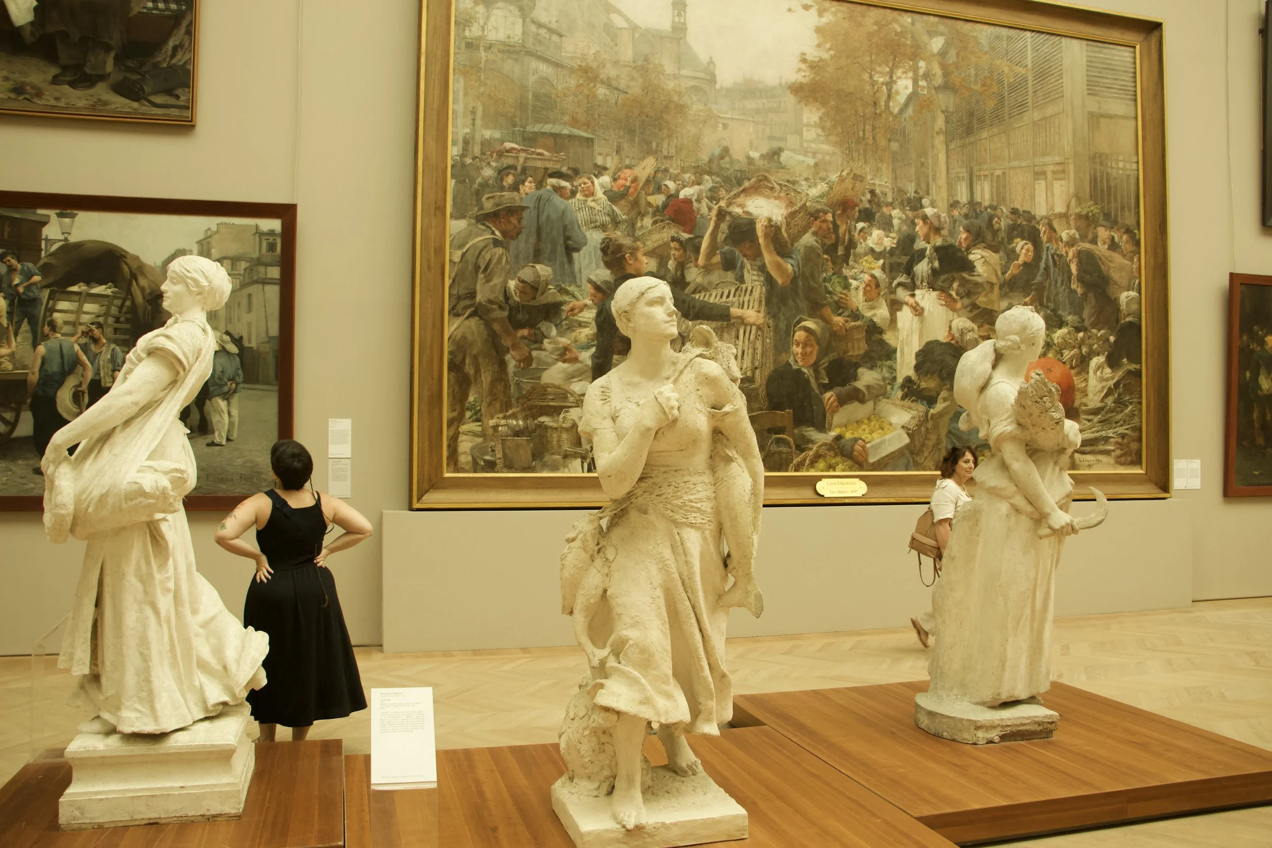 Museum gallery displaying white sculptures and a large framed painting of a crowded Paris market scene, with visitors viewing the artwork.