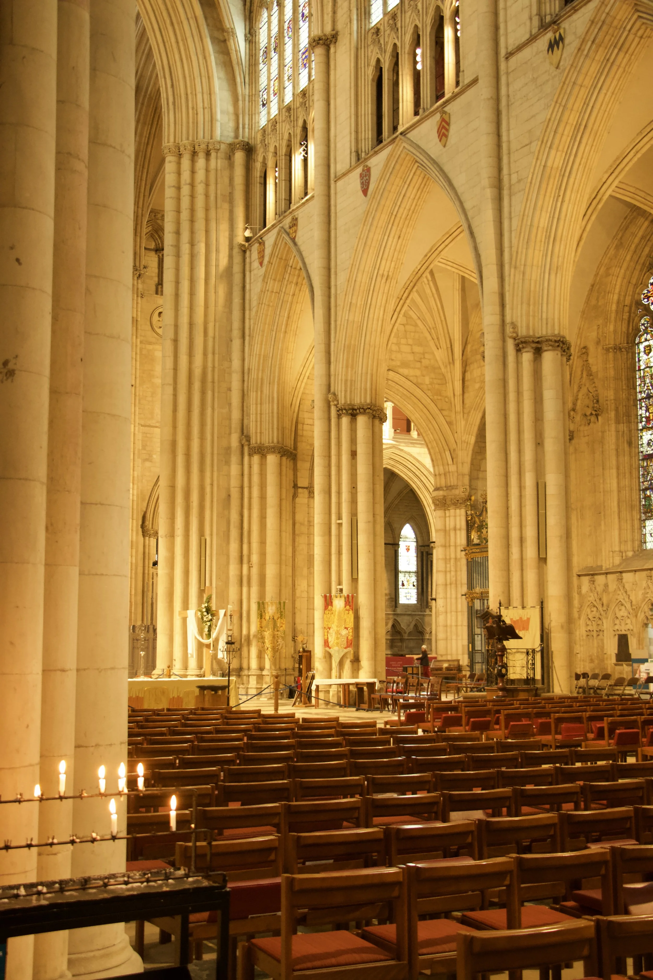 Interior of a Gothic cathedral with pointed arches, stained-glass windows, stone columns, church pews, candles, and an altar.