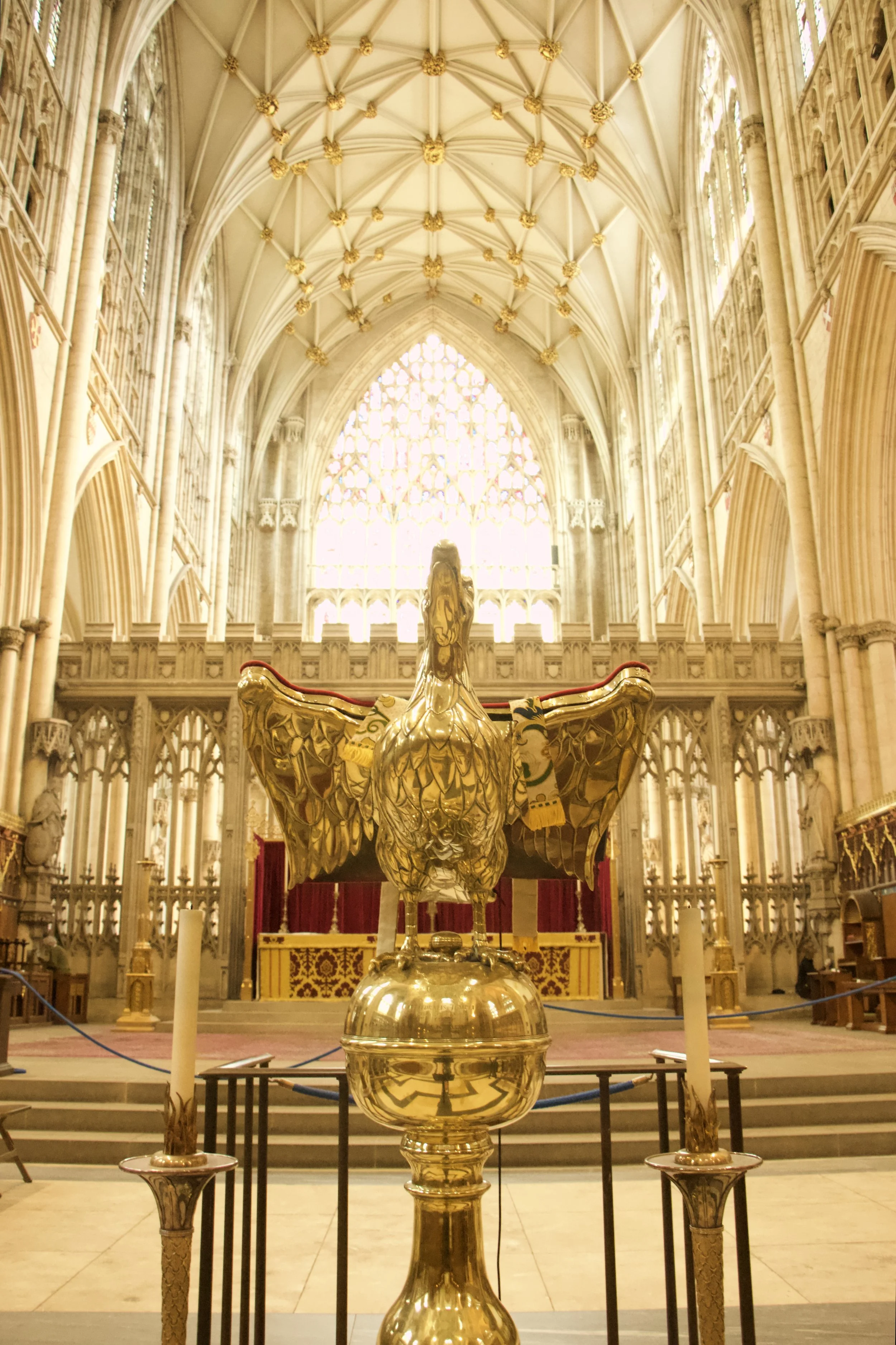 Ornate golden eagle lectern inside a York Minster's Gothic cathedral, with a vaulted ceiling, stained-glass window, altar, and candles.