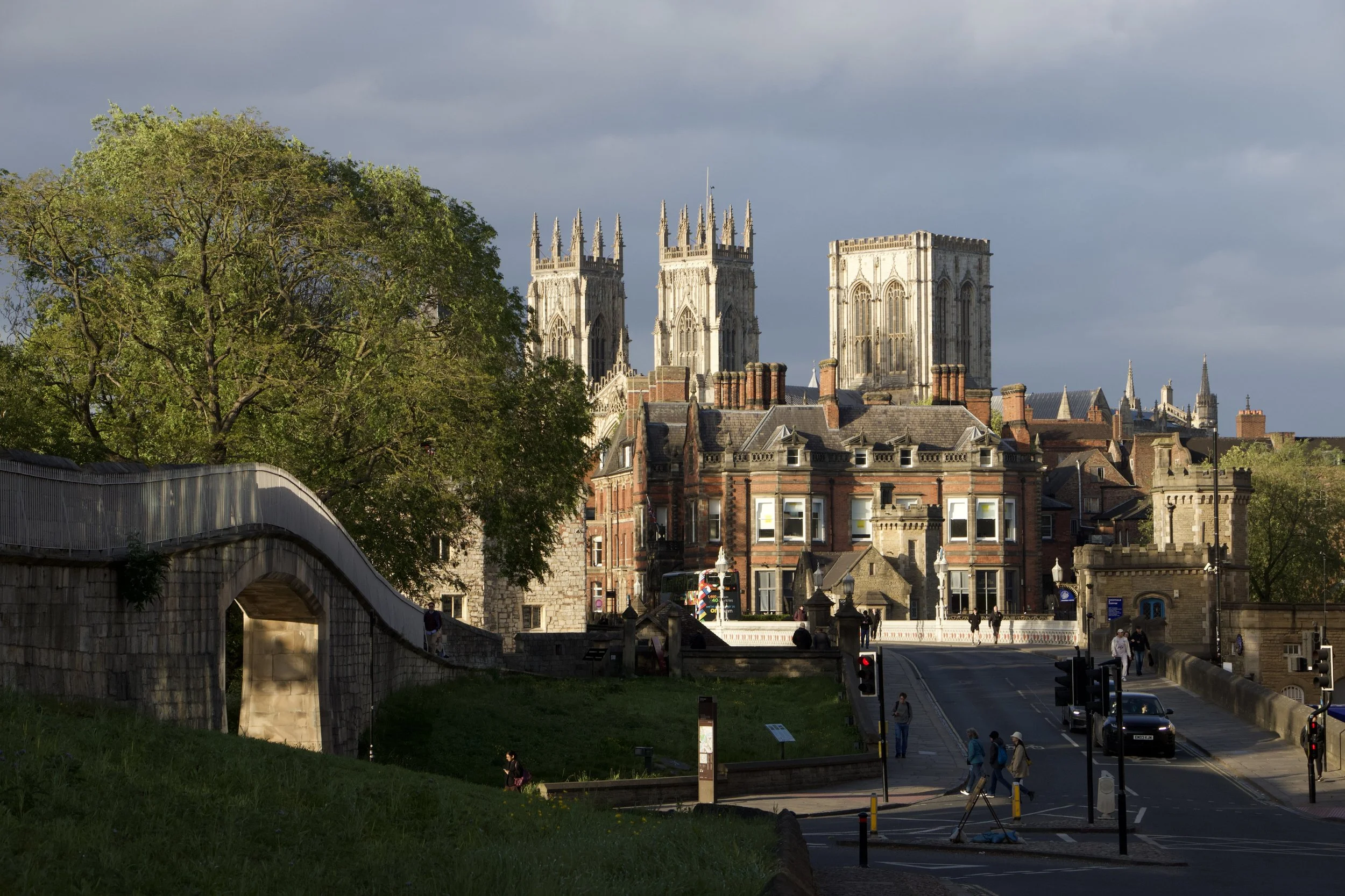 York Minster cathedral towers overlooking historic York, England, with a city street, stone bridge, trees, and pedestrians.