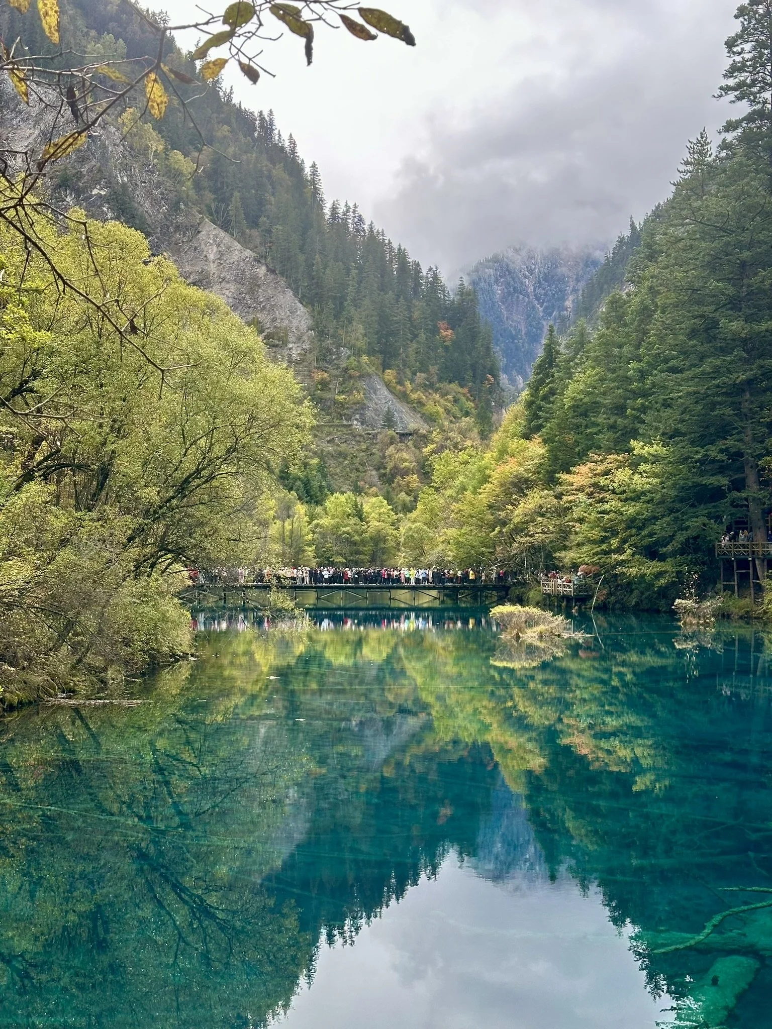 Visitors on a footbridge above a crystal-clear turquoise lake surrounded by forested mountains and autumn foliage in Jiuzhiagou National Park. 