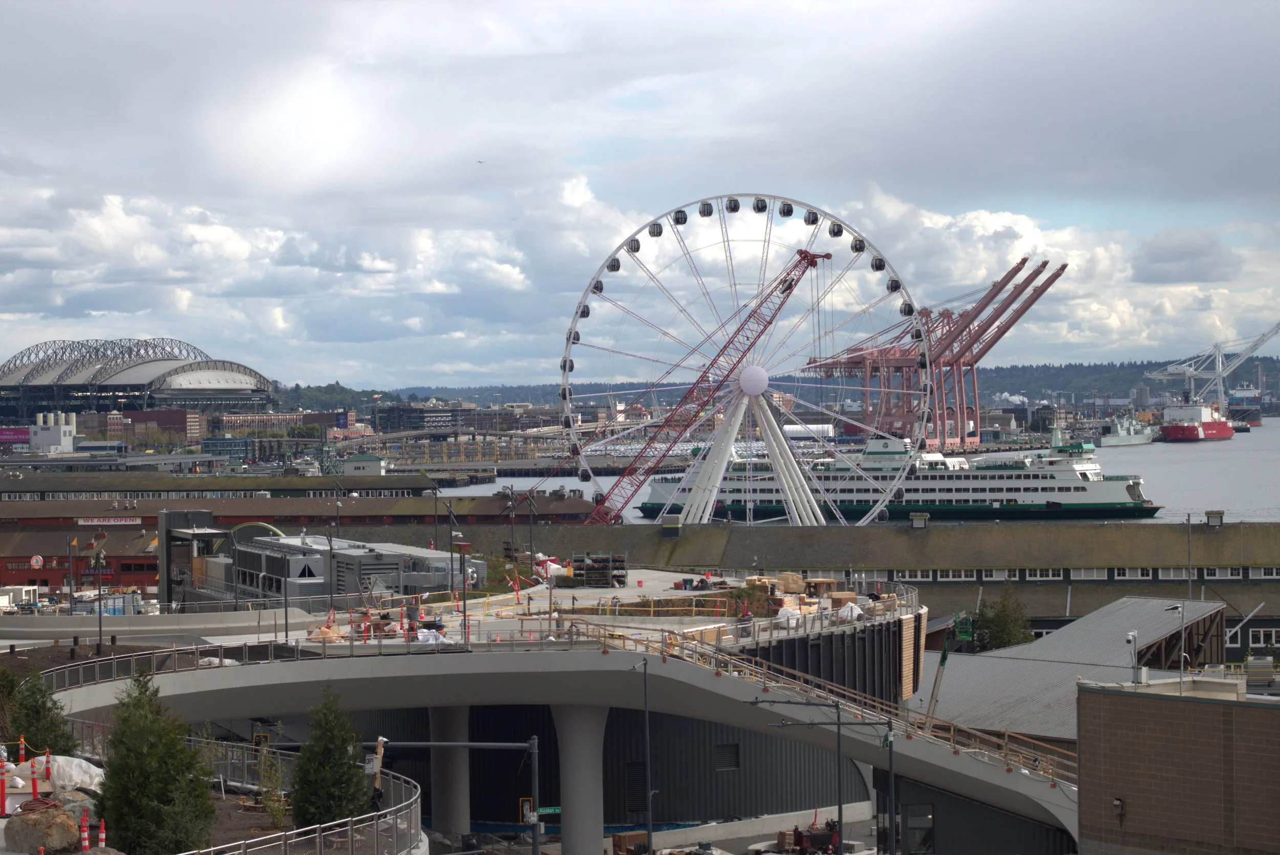 Seattle waterfront view with the Seattle Great Wheel, ferry, shipping cranes, and Lumen Field under a cloudy sky