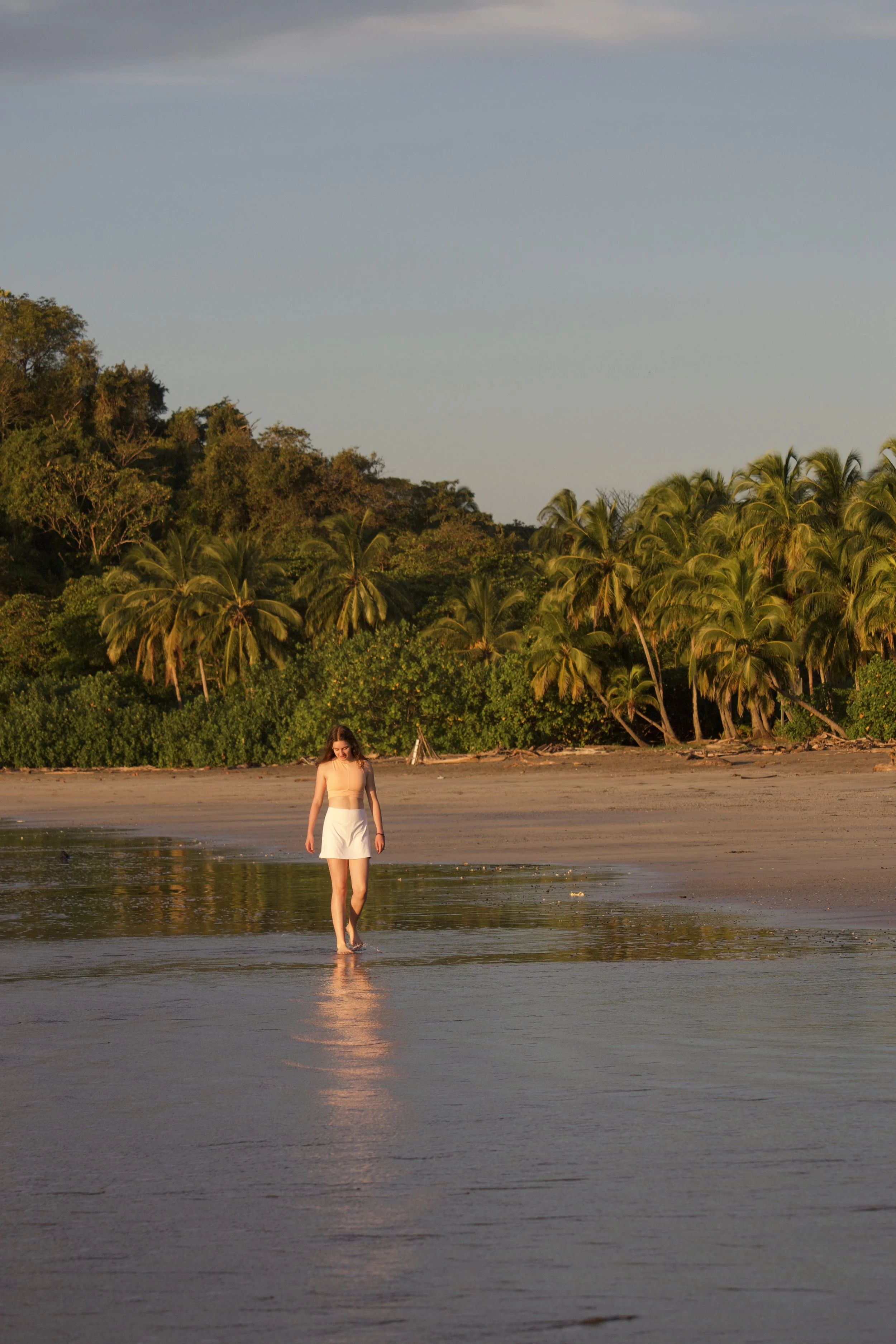 Woman walking barefoot through shallow water on a tropical beach at sunset, with palm trees in the background