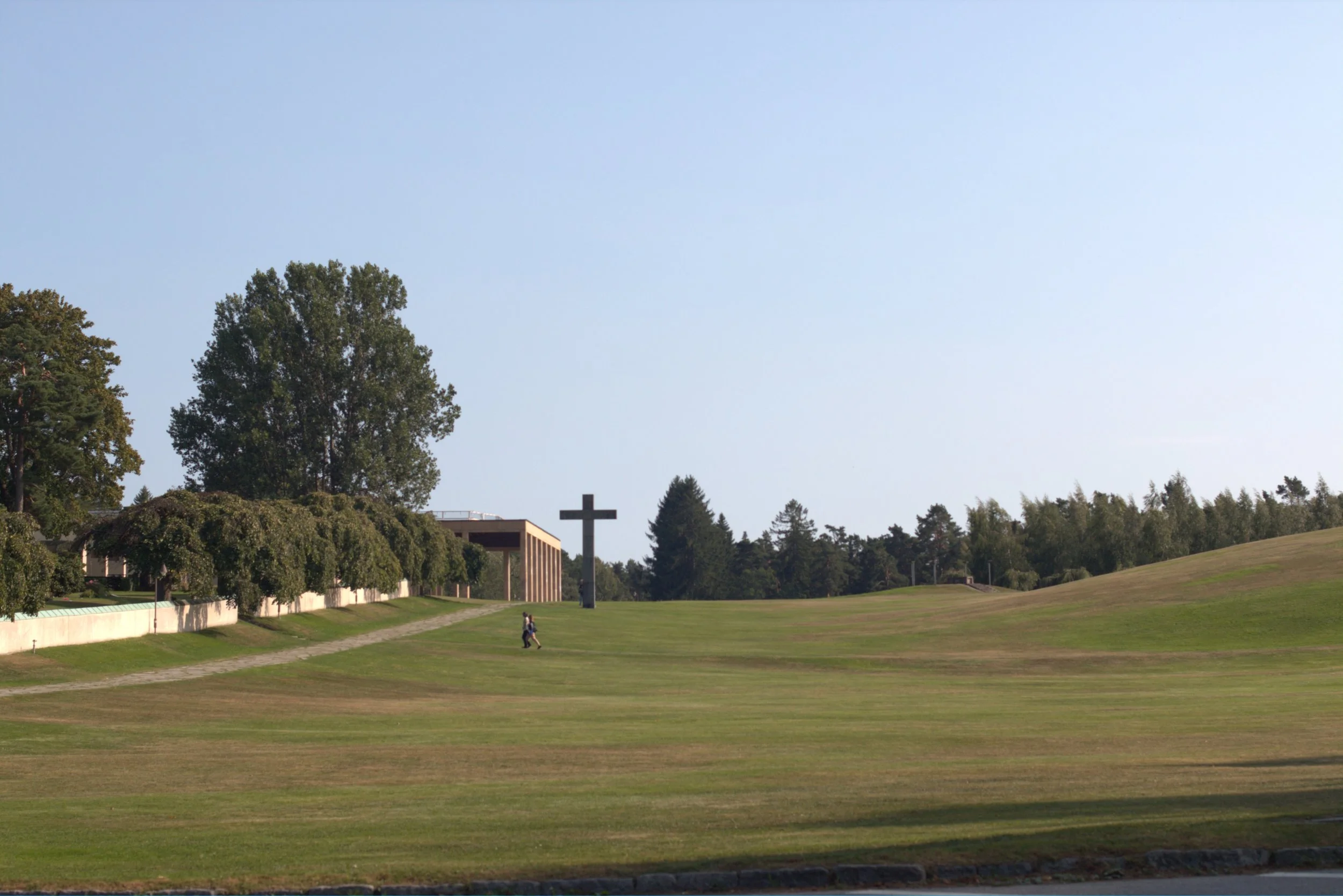 Large cross monument beside a modern building in a landscaped grassy park with trees and a person walking