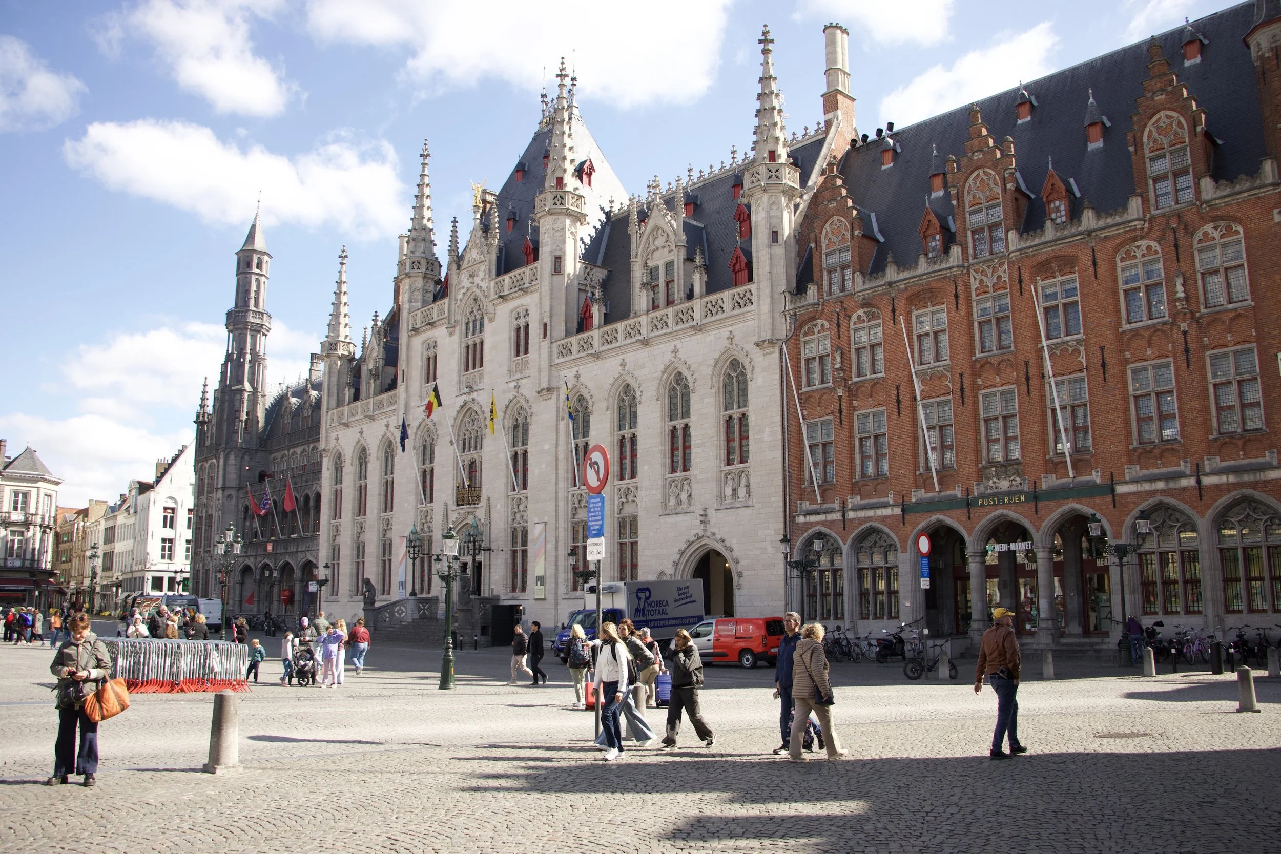 Historic Gothic buildings and pedestrians in Bruges, Belgium, under a bright blue sky.