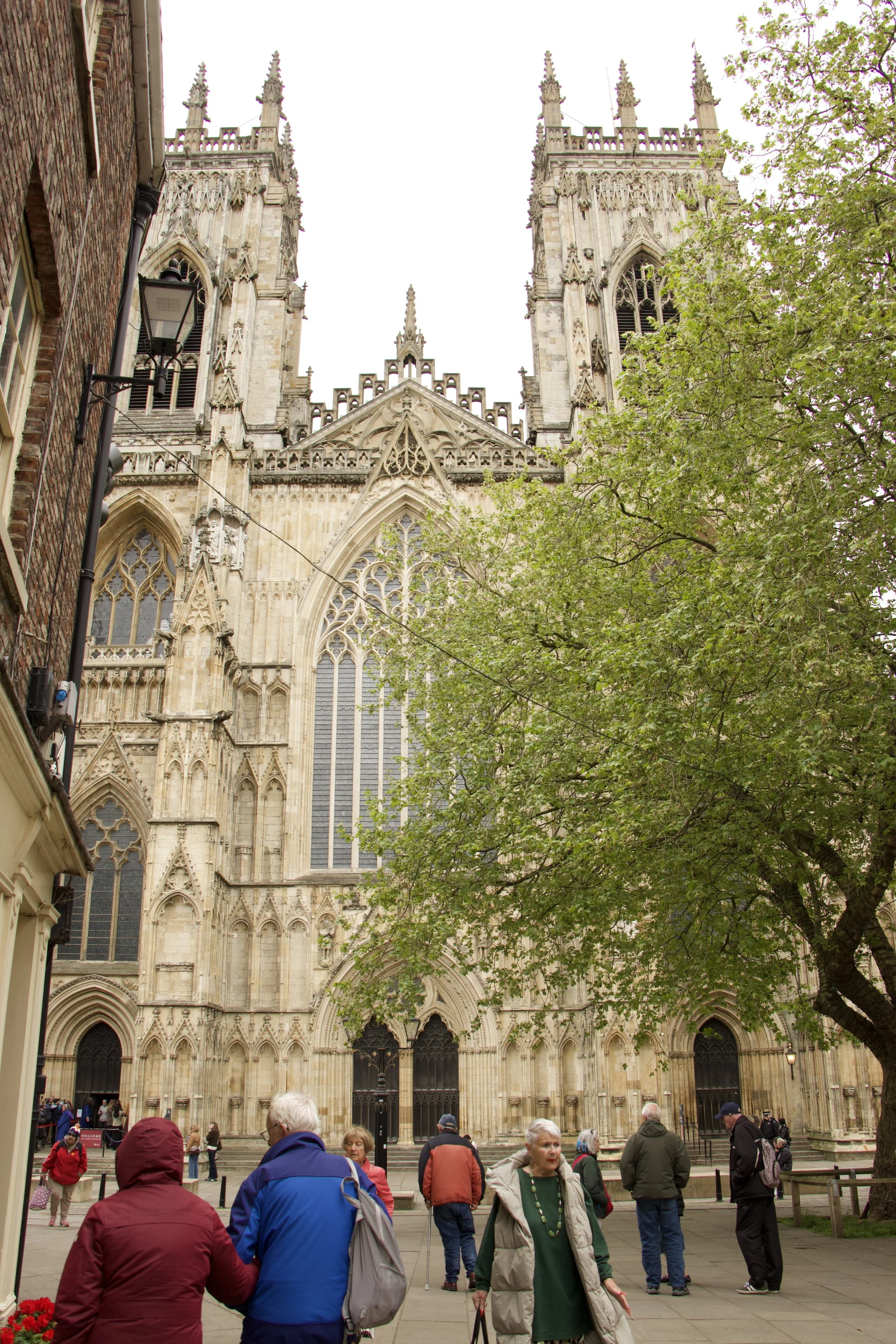 Visitors in a cathedral courtyard viewing an ornate Gothic cathedral facade with pointed towers, arched windows, and tree branches.