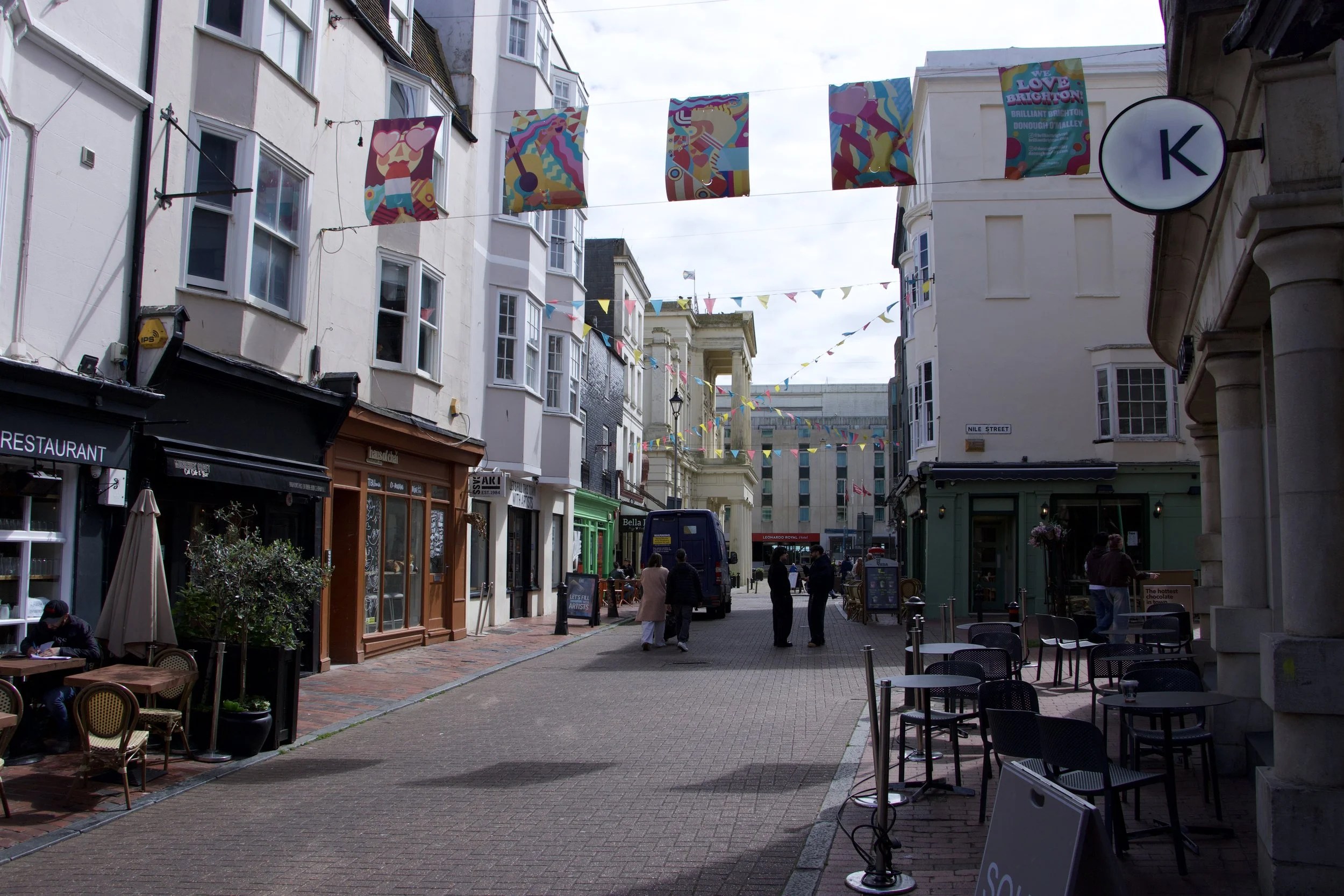 Pedestrian street in Brighton, England, lined with restaurants and cafes, colorful bunting, and hanging art banners.