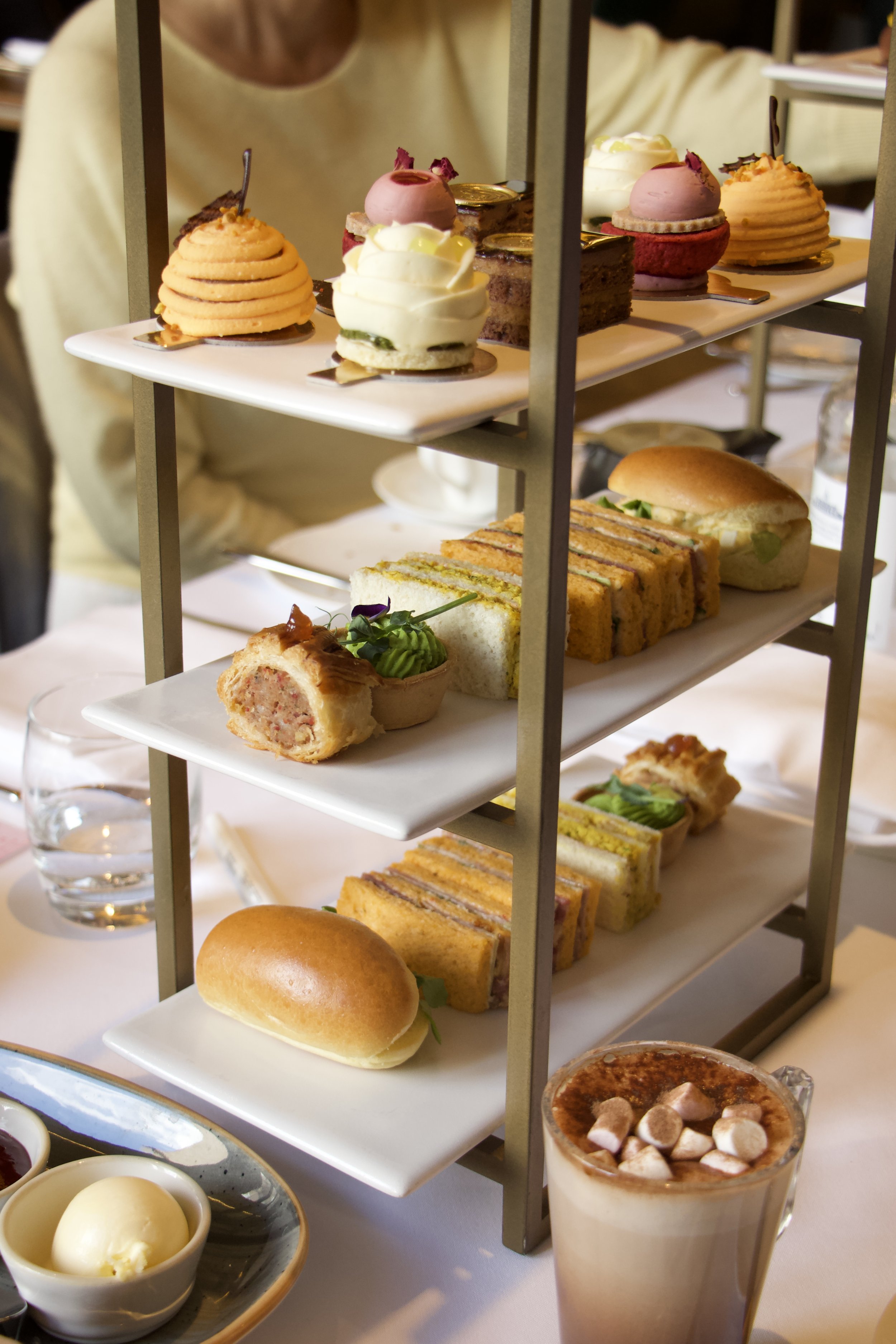 Tiered afternoon tea stand with assorted pastries, sandwiches, desserts, coffee, and hot chocolate topped with marshmallows