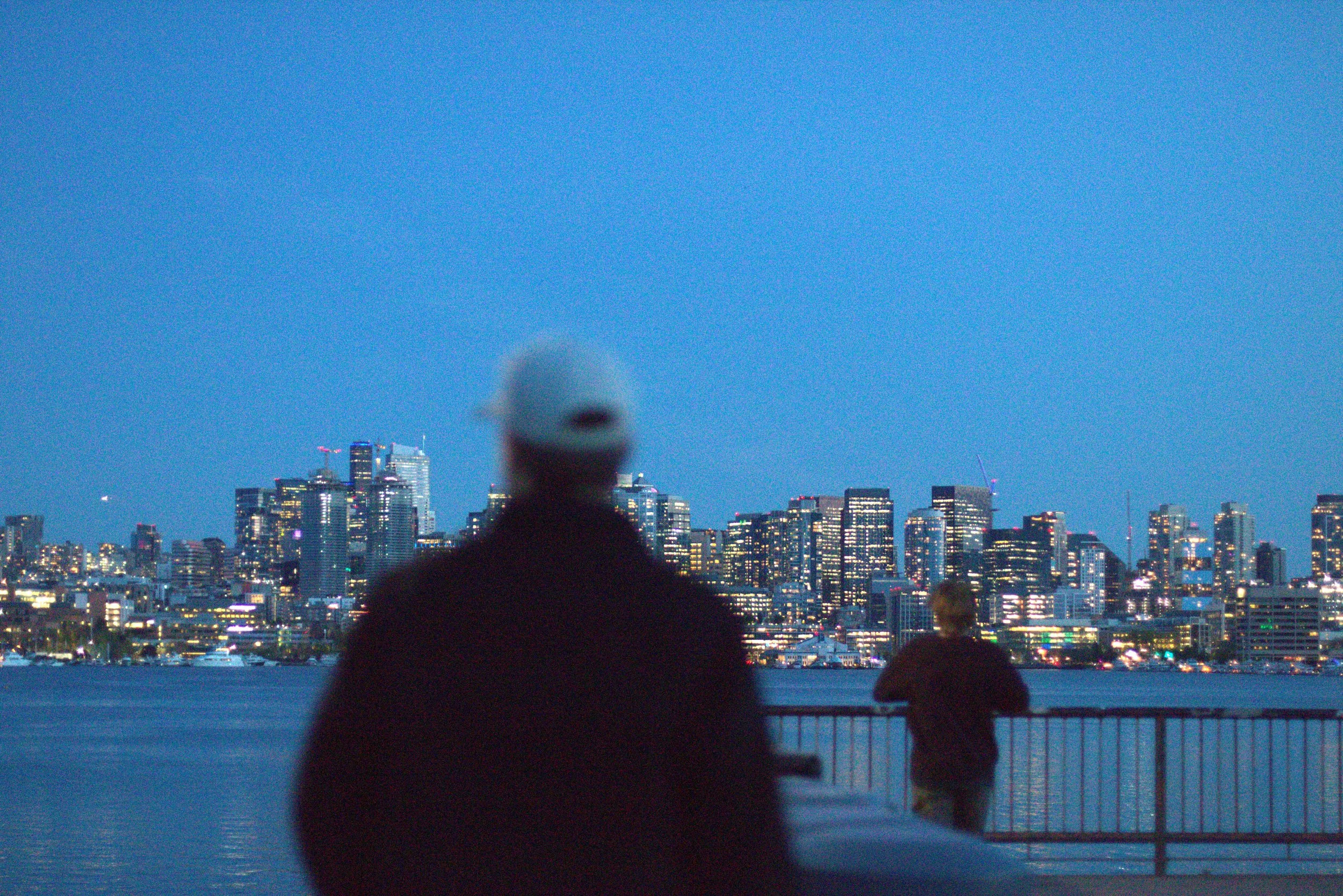 Blurred people overlooking a brightly lit city skyline across the water at dusk