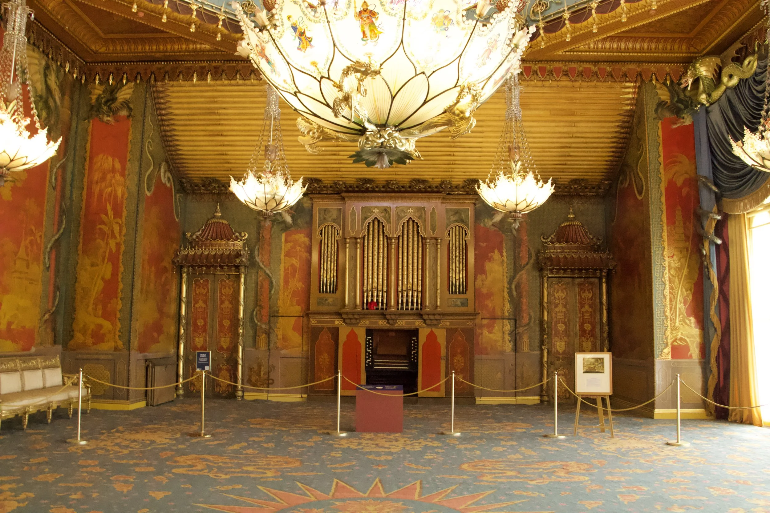 Ornate music room interior with a central pipe organ, illuminated lotus-shaped chandeliers, gilded woodwork, and colorful decorative murals.