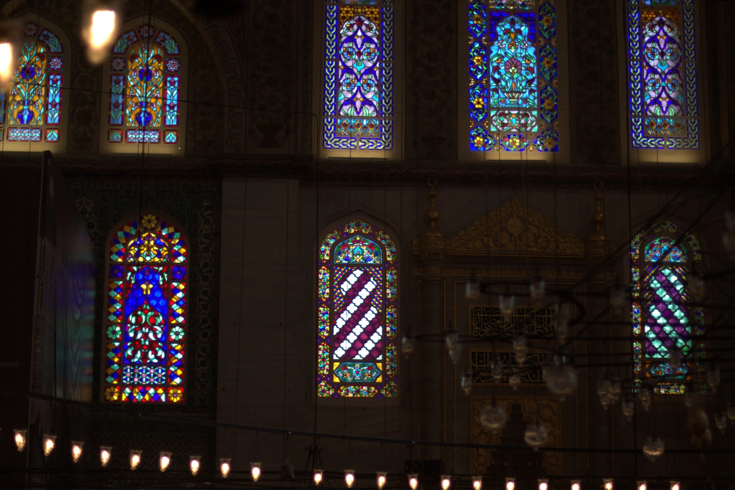 Interior of a mosque with ornate colorful stained-glass windows, chandeliers, and Arabic calligraphy.