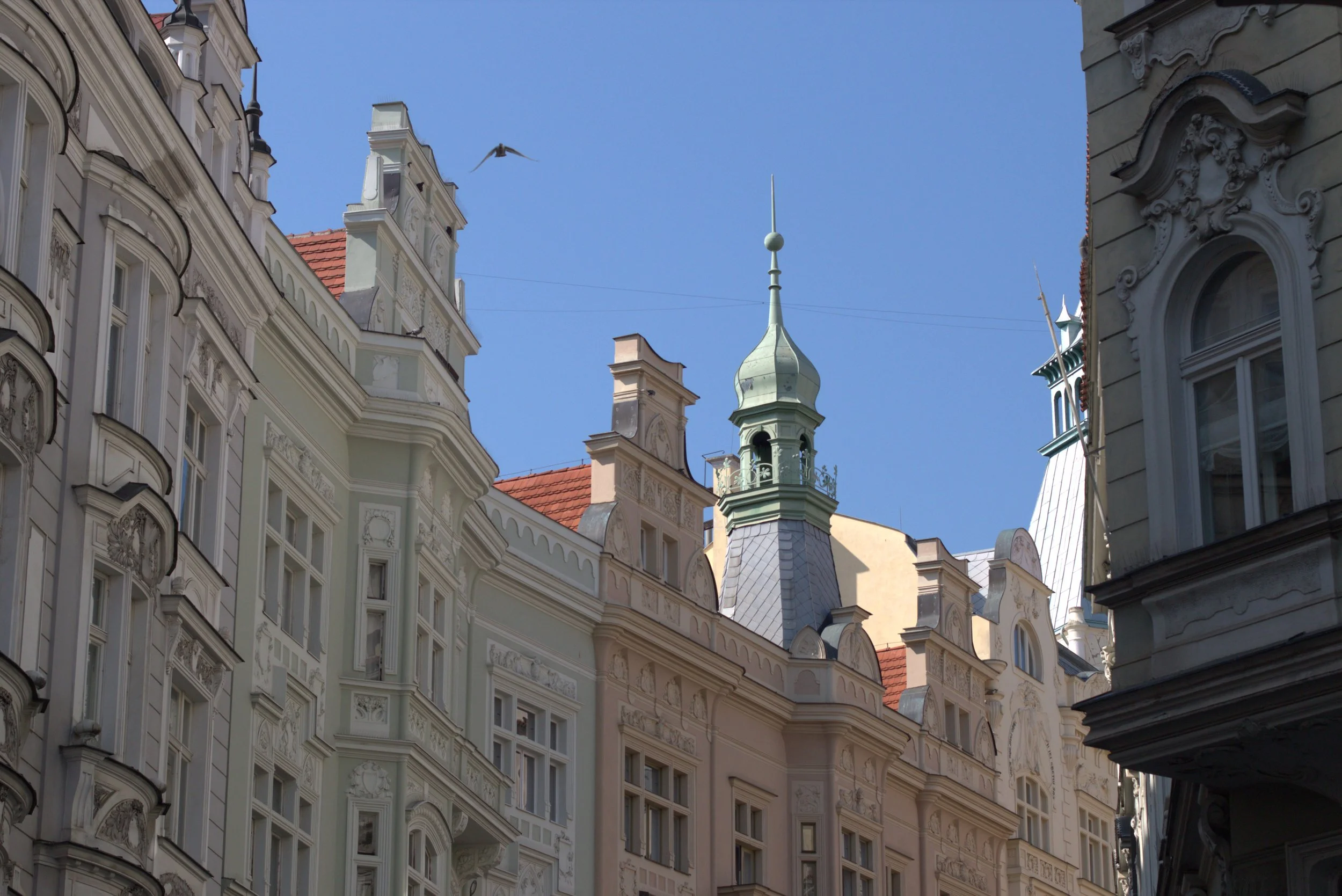 Ornate historic European buildings with decorative facades, red tile roofs, and a green domed turret under a clear blue sky