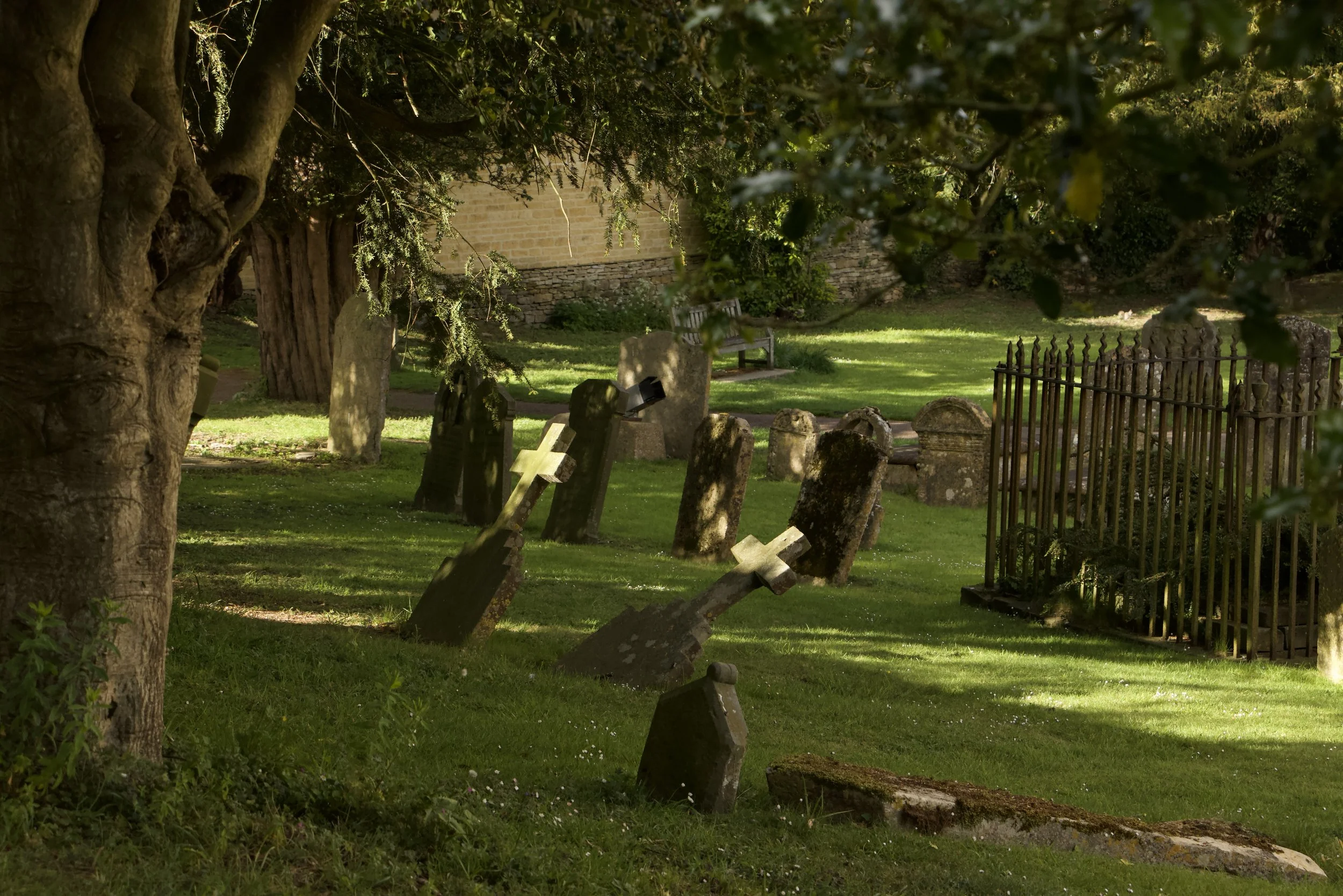 The sunlit Blockley cemetery with weathered gravestones, trees, stone wall, and wrought-iron grave enclosure.