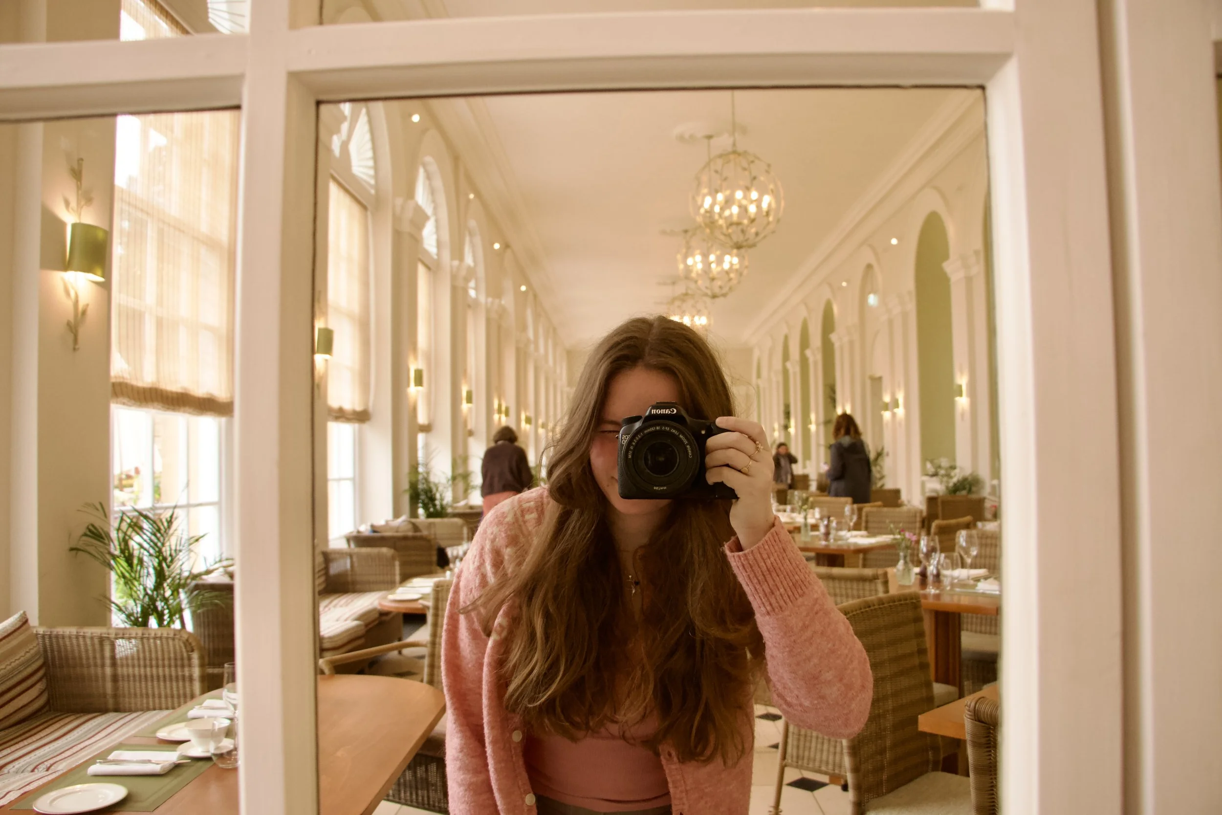 Woman in a pink sweater taking a mirror selfie with a Canon camera in an elegant restaurant dining room