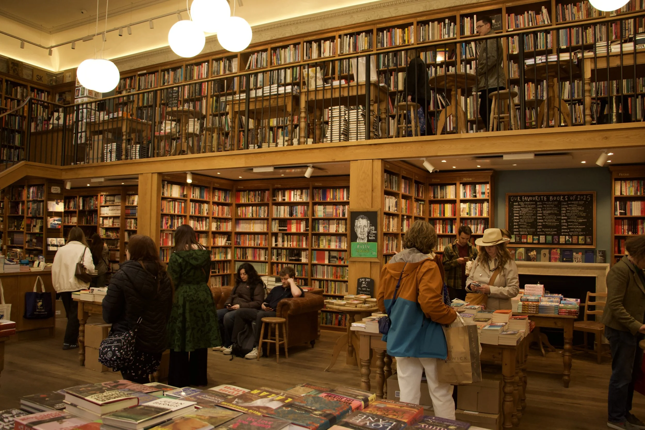 Customers browsing books inside a crowded independent bookstore with floor-to-ceiling shelves, a mezzanine, and warm  lighting.