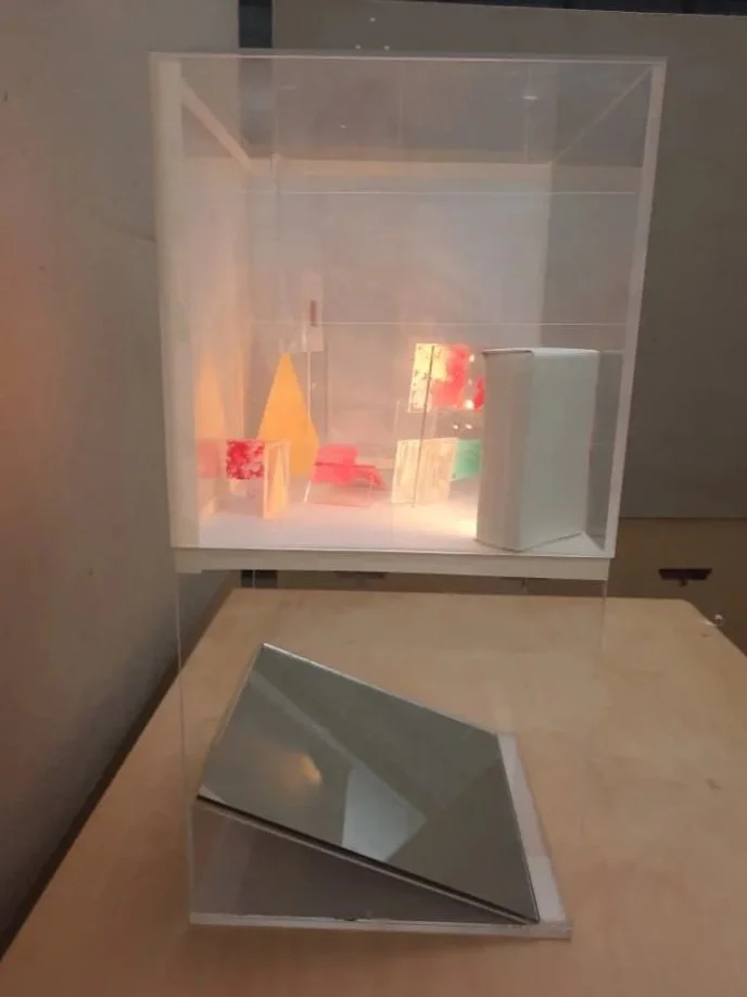 Illuminated handmade plexiglass display case half covered in tracing paper filled with colorful abstract geometric artwork completed by an angled reflective mirror on a table.