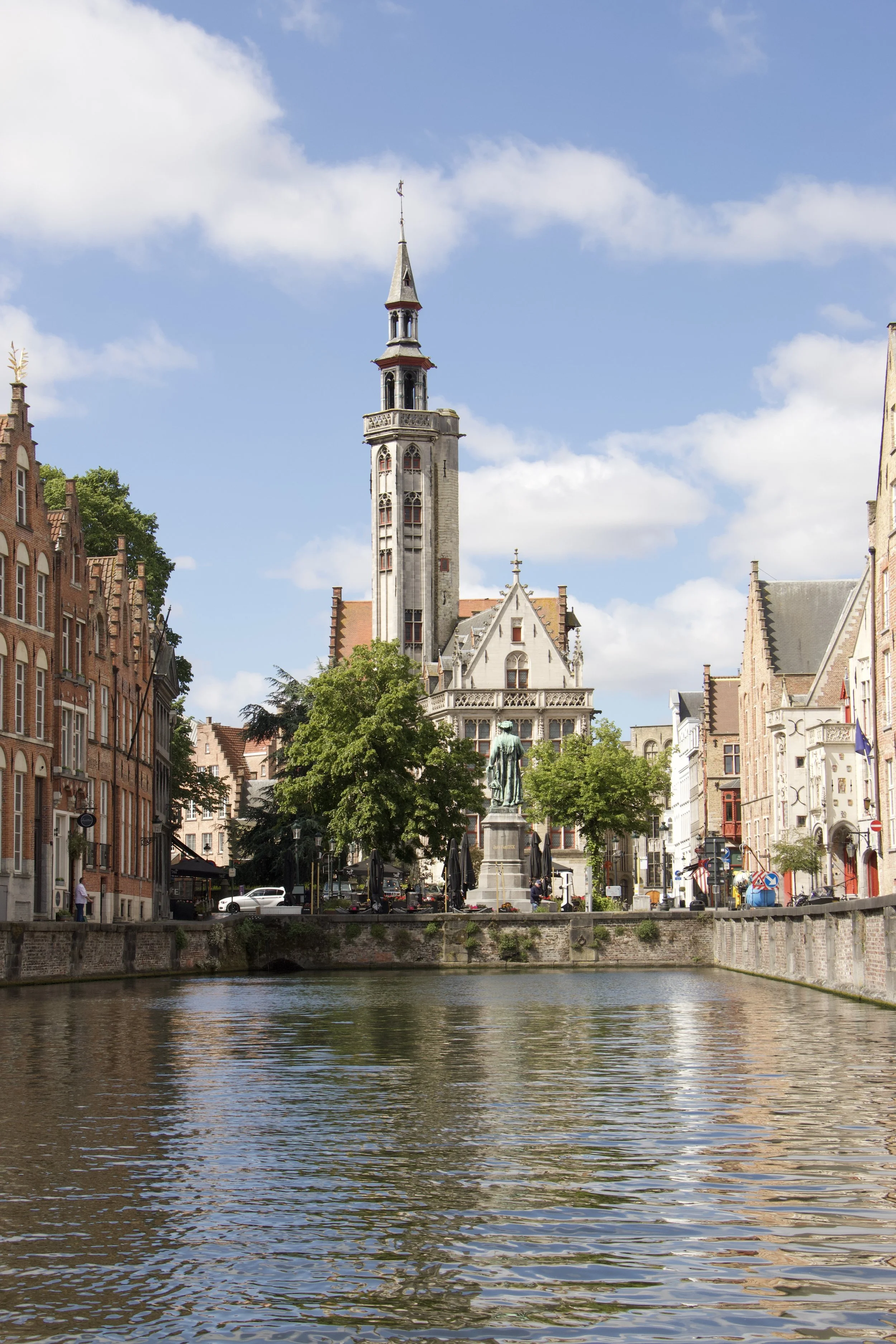 Historic Bruges, Belgium cityscape with a Gothic church tower, statue, canal, and waterfront buildings under a blue sky.