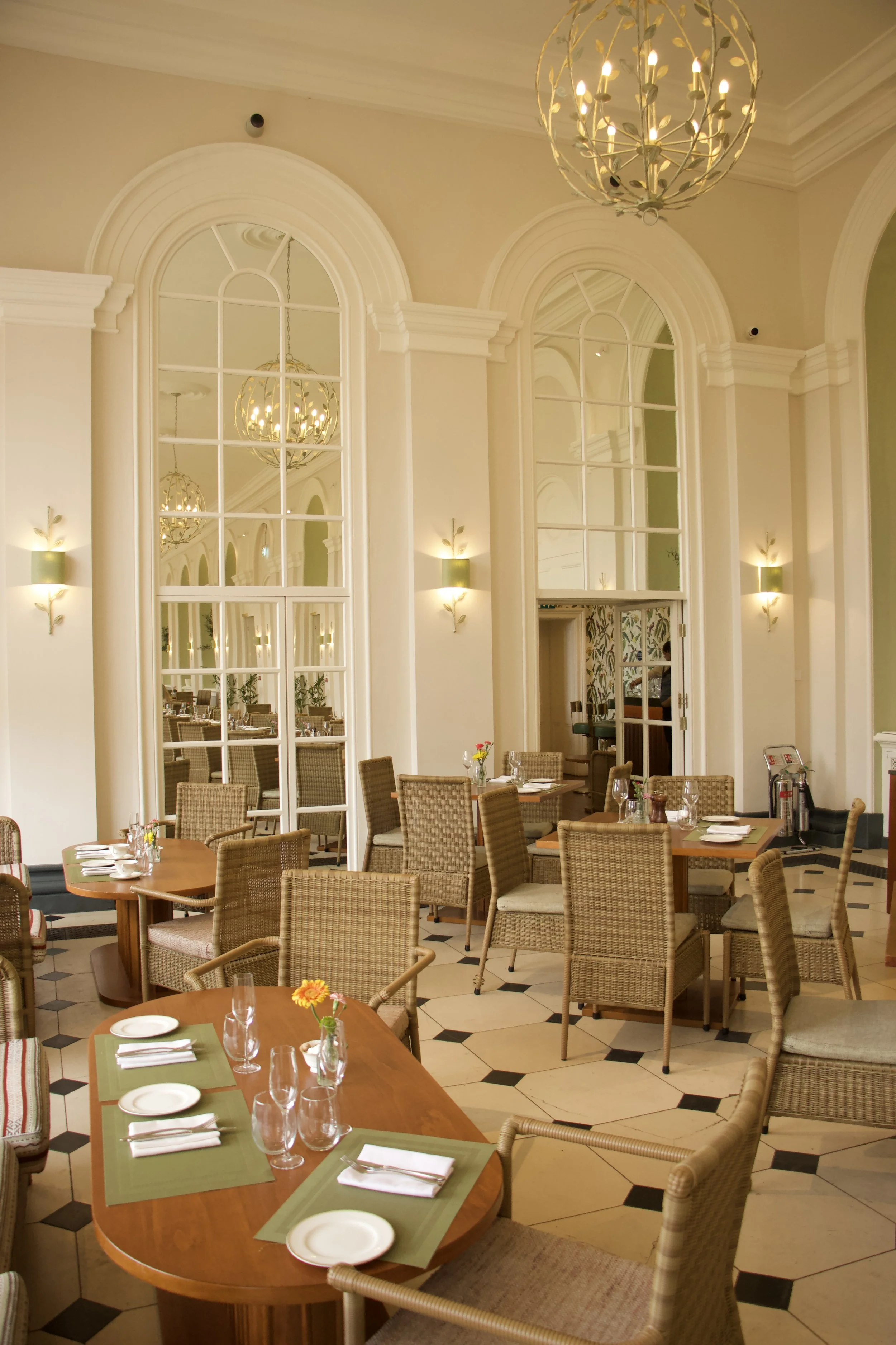 Elegant restaurant dining room in Blenheim Palace with wicker chairs, set tables, arched mirrors, and chandeliers.