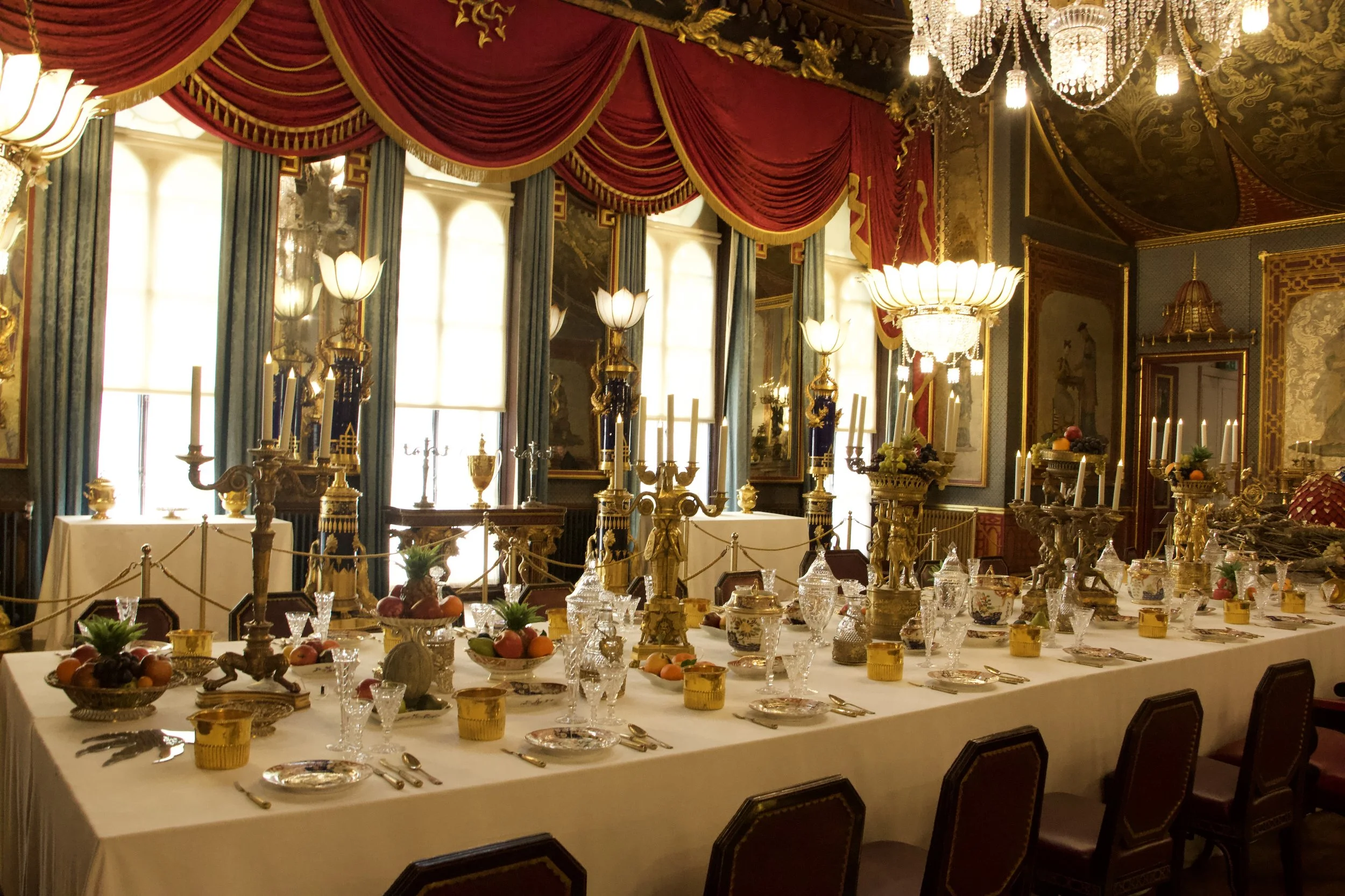 Ornate palace dining room with a long banquet table, crystal glassware, fruit centerpieces, gold candelabras, chandeliers, and red velvet drapery. 