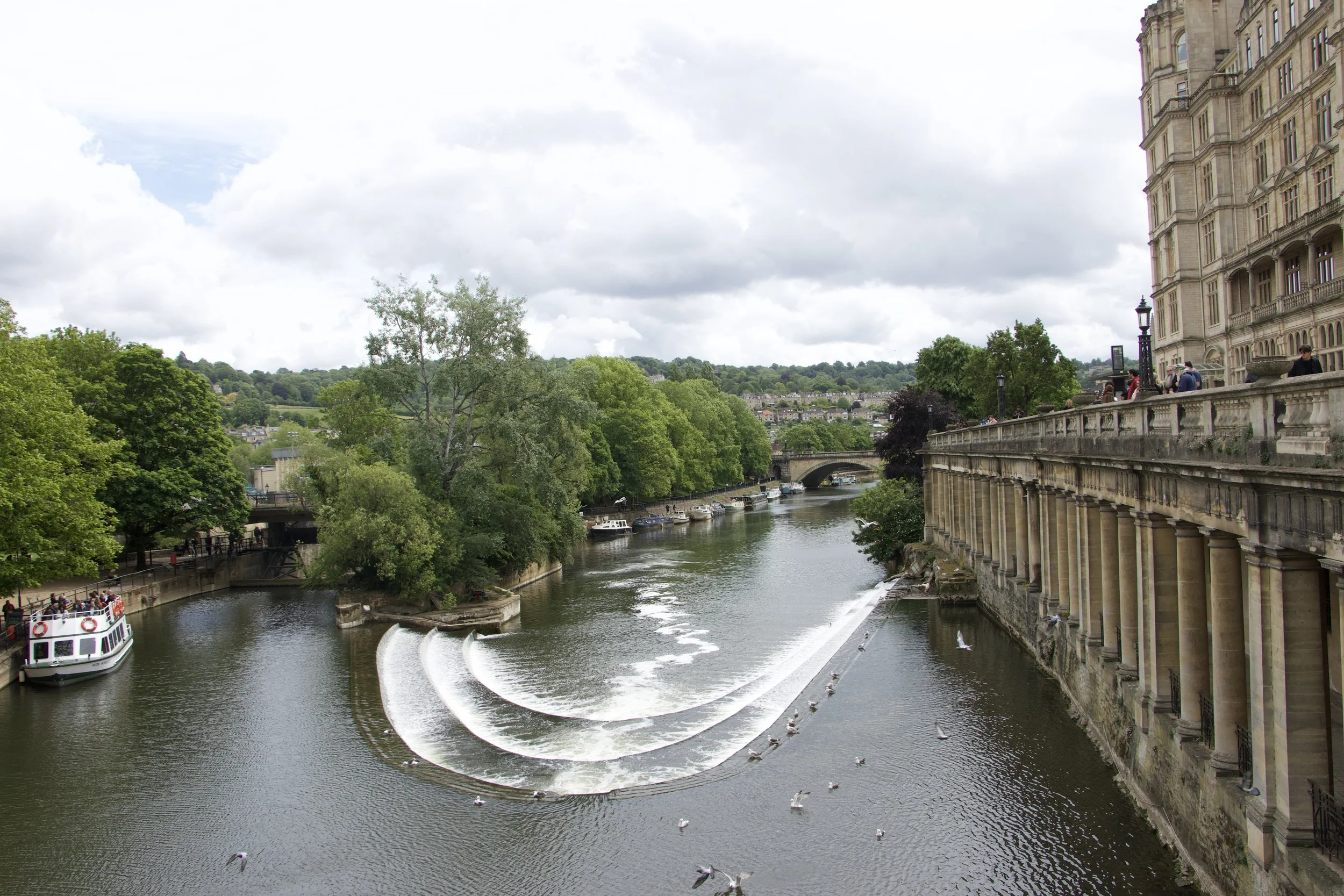 Panoramic view of the River Avon and Pulteney Weir in Bath, England, with a historic stone building, boats, and hillside greenery.
