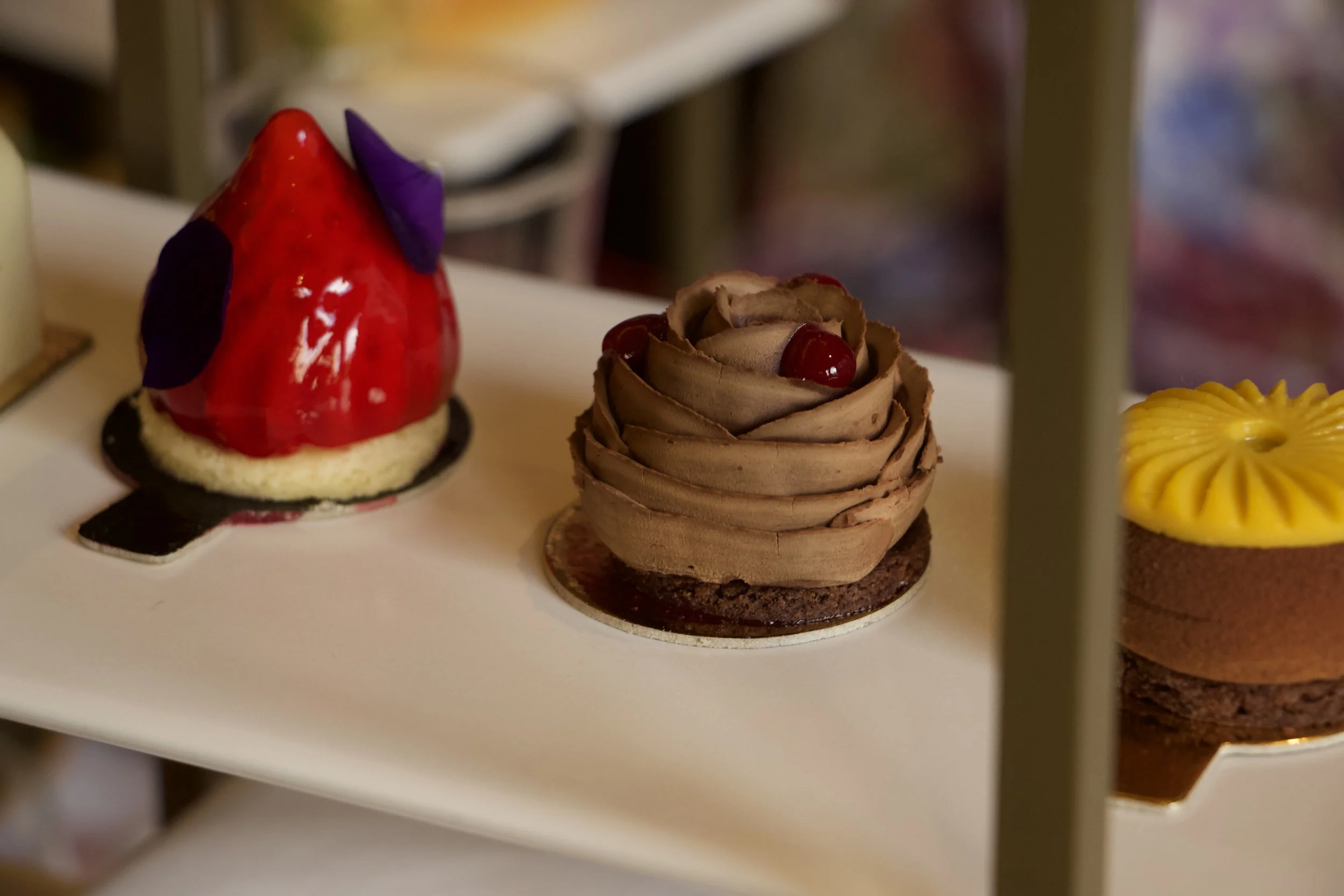 Assorted gourmet pastries, including a red and purple cone-shaped tart, a chocolate layered rose cake with cherries, and a yellow-topped dessert.