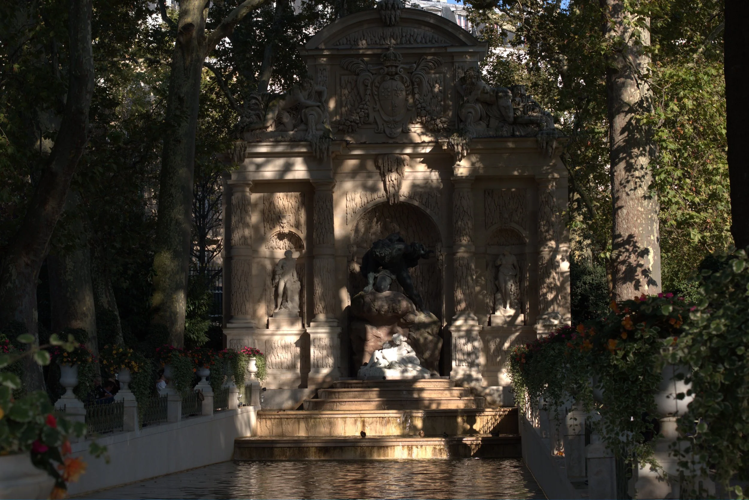 Ornate stone fountain with sculptures, coat of arms, and cascading pools in a tree-lined garden