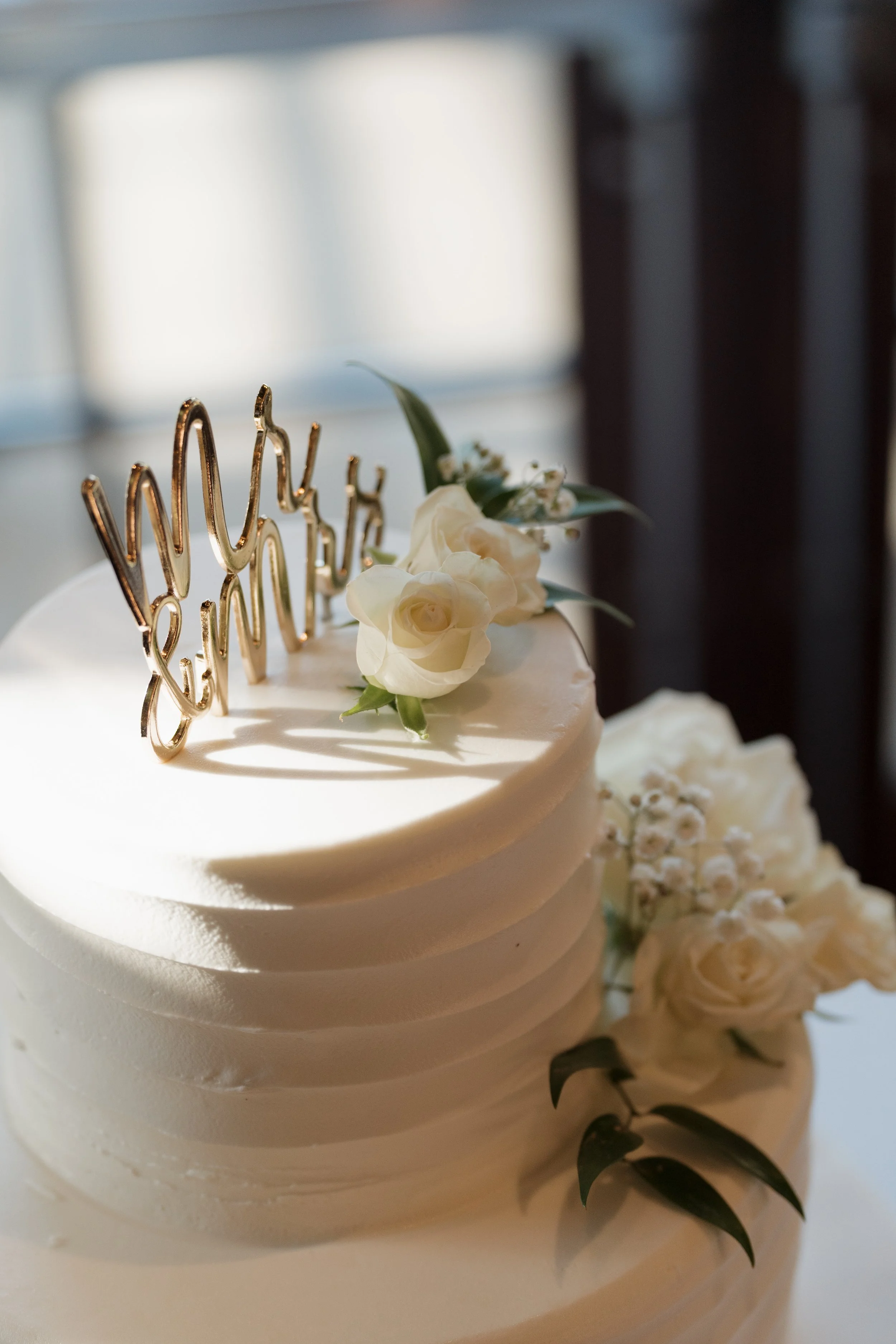 Close-up of a white wedding cake decorated with white roses, greenery, and a gold script cake topper.