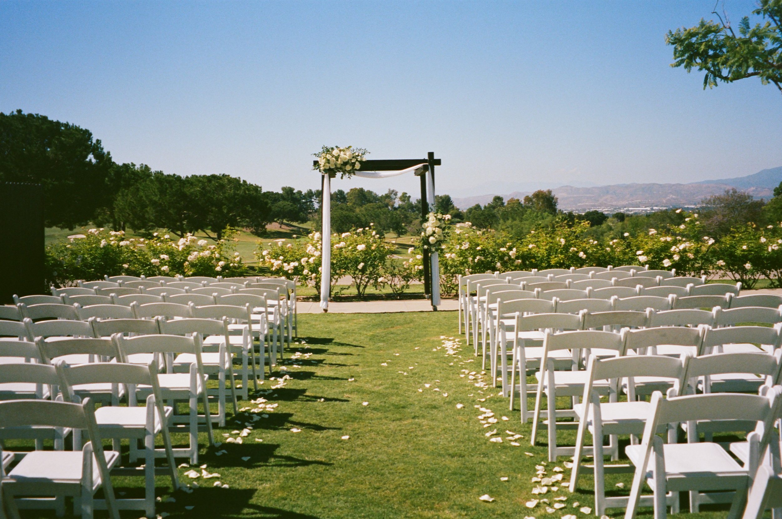 Outdoor wedding ceremony setup with white chairs, flower-adorned wooden arch, rose garden, and mountain view