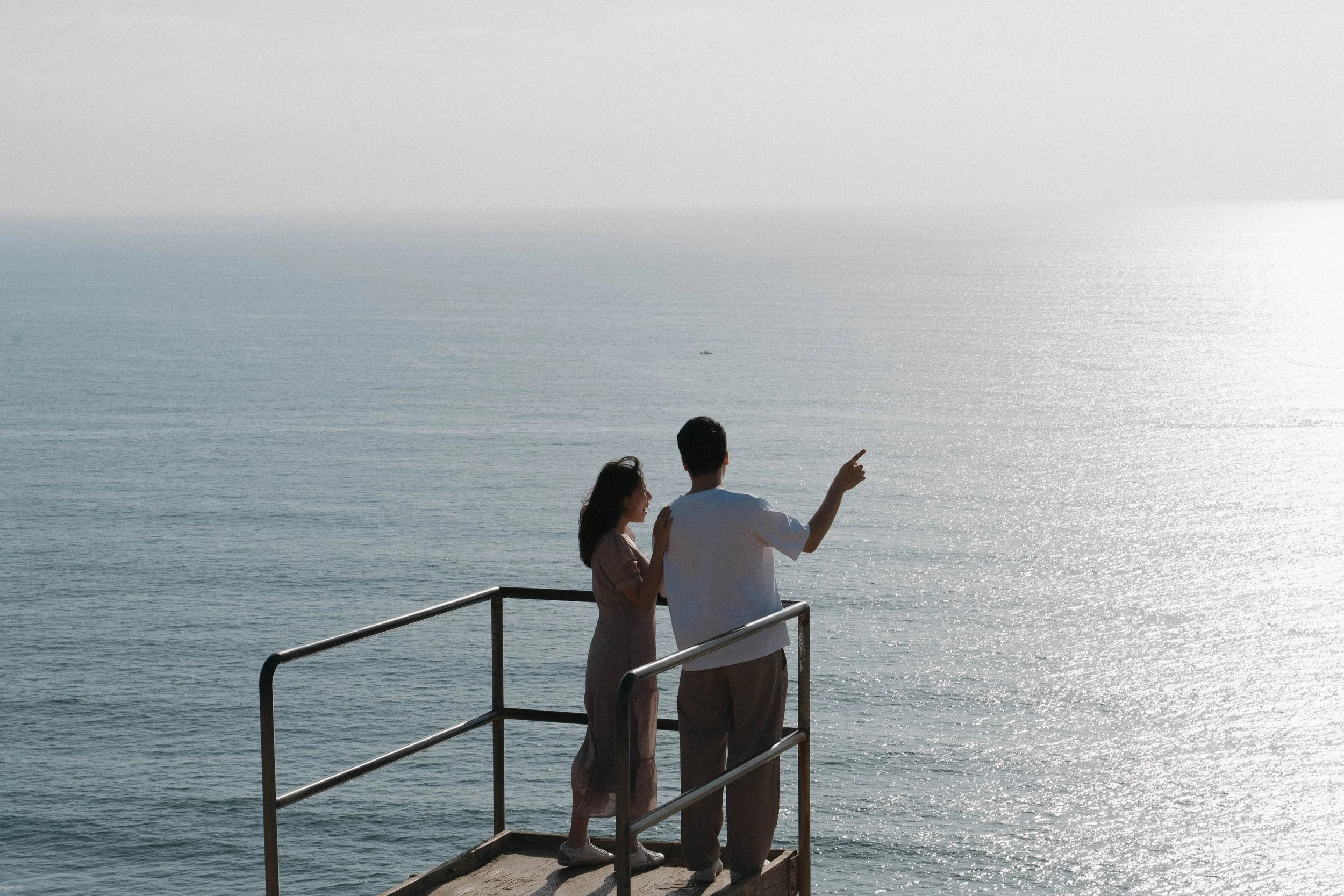 Couple standing on a seaside viewing platform overlooking a sunlit ocean