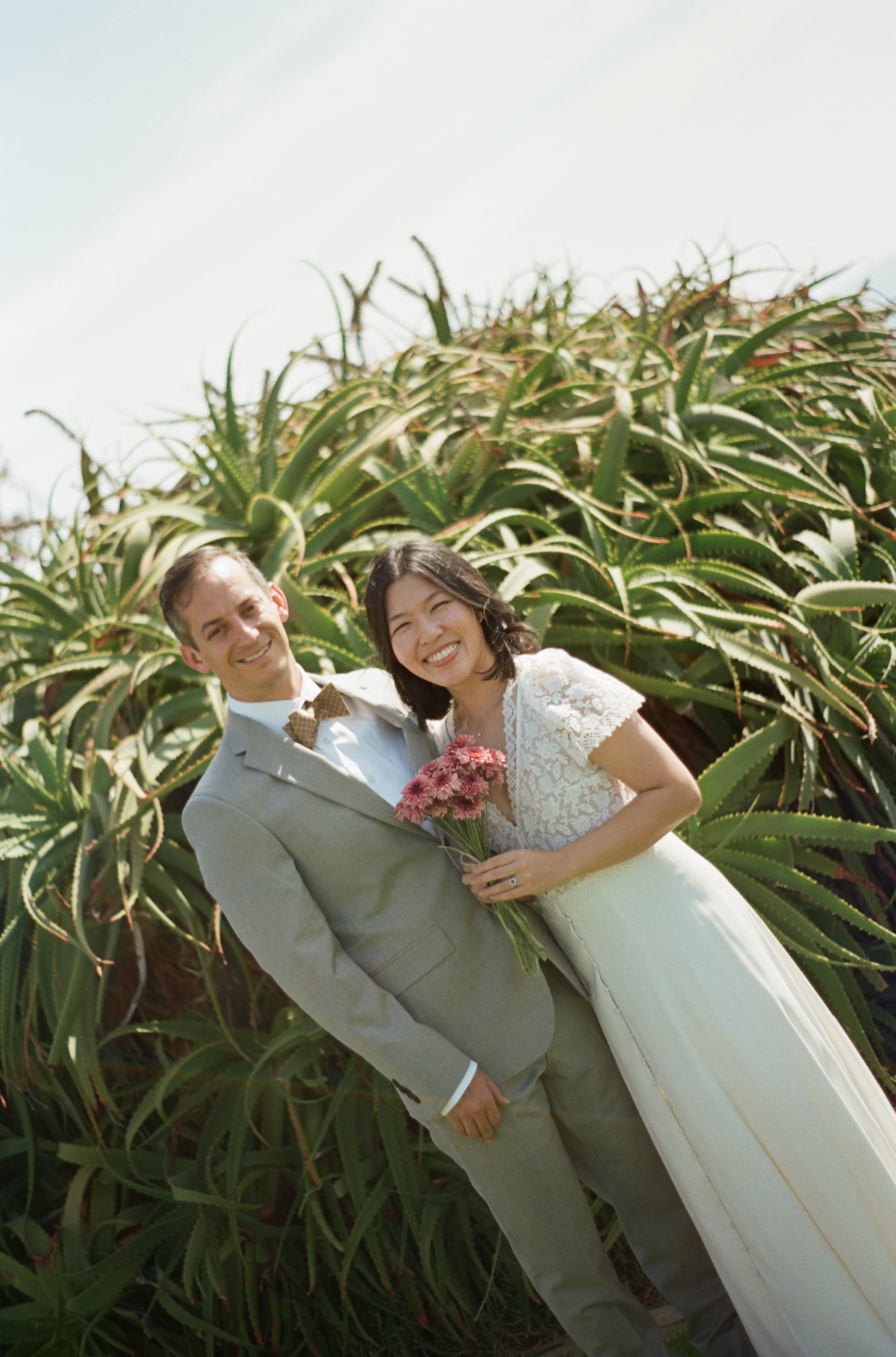 Newlywed couple smiling in wedding attire while holding flowers in front of aloe plants