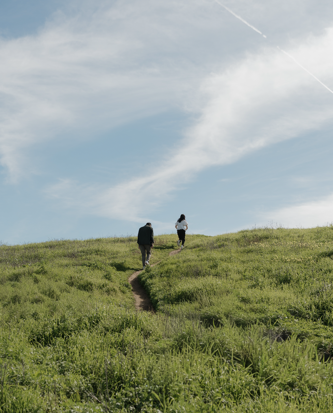 Two people hiking along a narrow trail through a green meadow beneath a blue, cloud-filled sky.