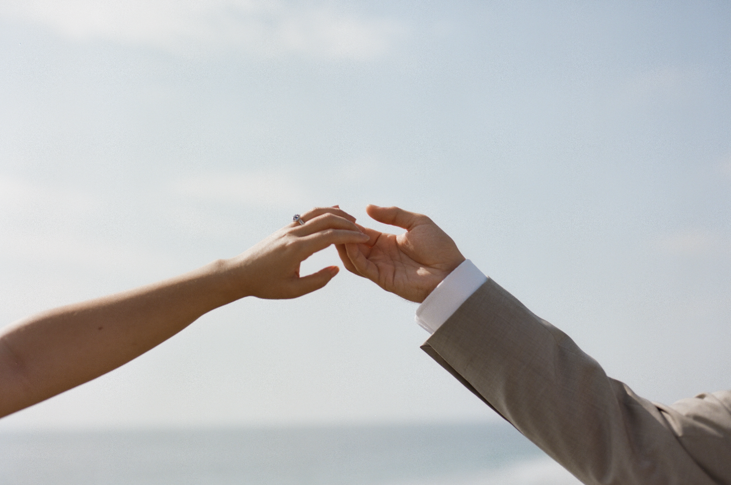 Two people reaching toward each other against a bright sky, one wearing a wedding ring and the other a suit.