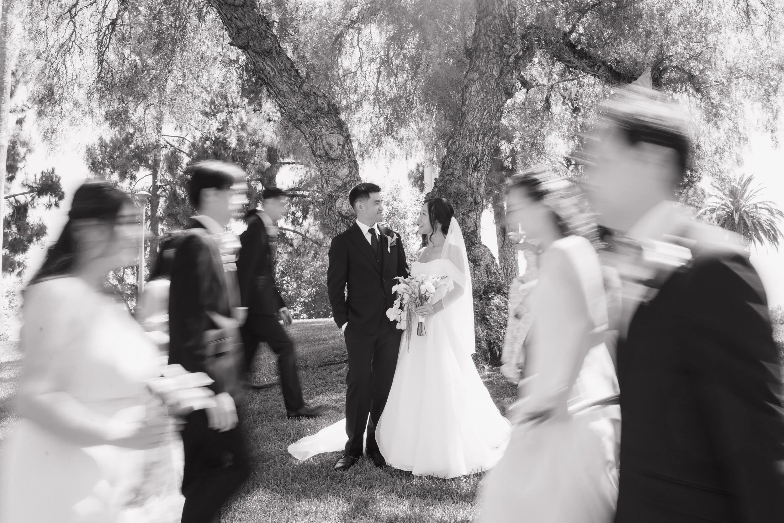 Black-and-white wedding portrait of a bride and groom standing beneath trees as blurred wedding guests walk around them.