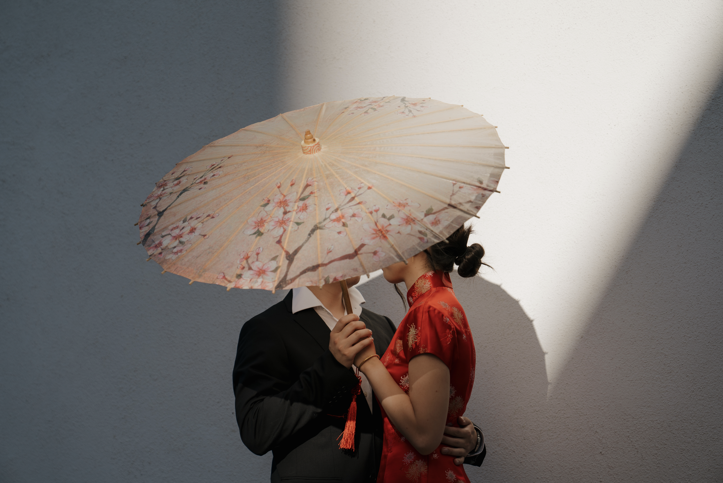 Couple embracing beneath a floral paper parasol, with one person wearing a red traditional Chinese dress and the other a black suit
