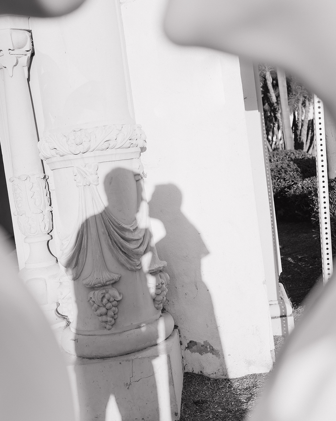 Black-and-white photo of an ornate classical column capital with floral carvings and draped stone details, beside a building wall.