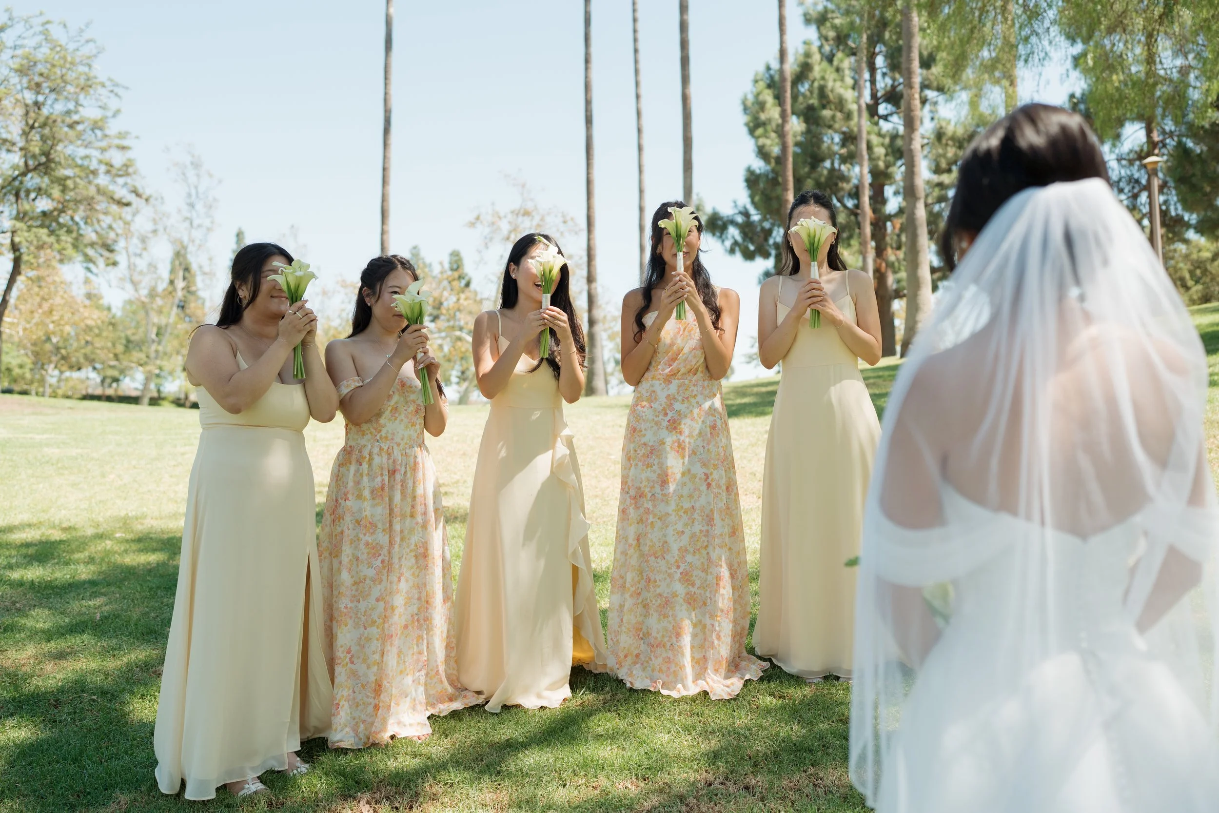 Bride in a white veil faces five bridesmaids holding calla lily bouquets at an outdoor wedding