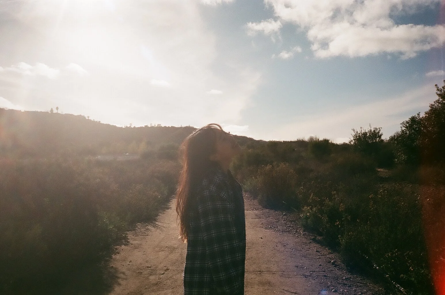 Backlit person with long hair walking along a dirt trail through a sunlit hillside landscape