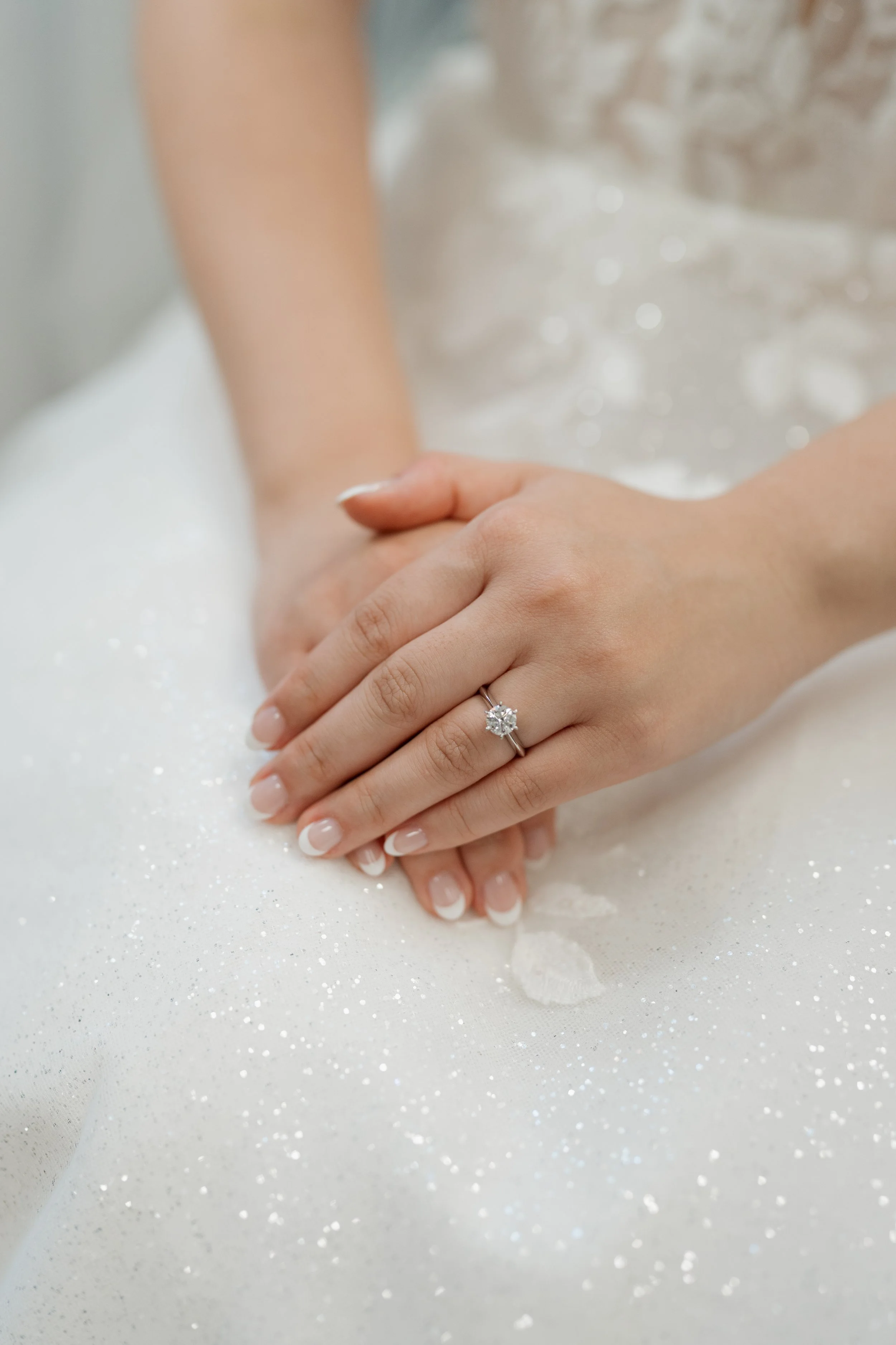 Close-up of a bride’s hands with French manicure and diamond engagement ring resting on a sparkly white wedding dress