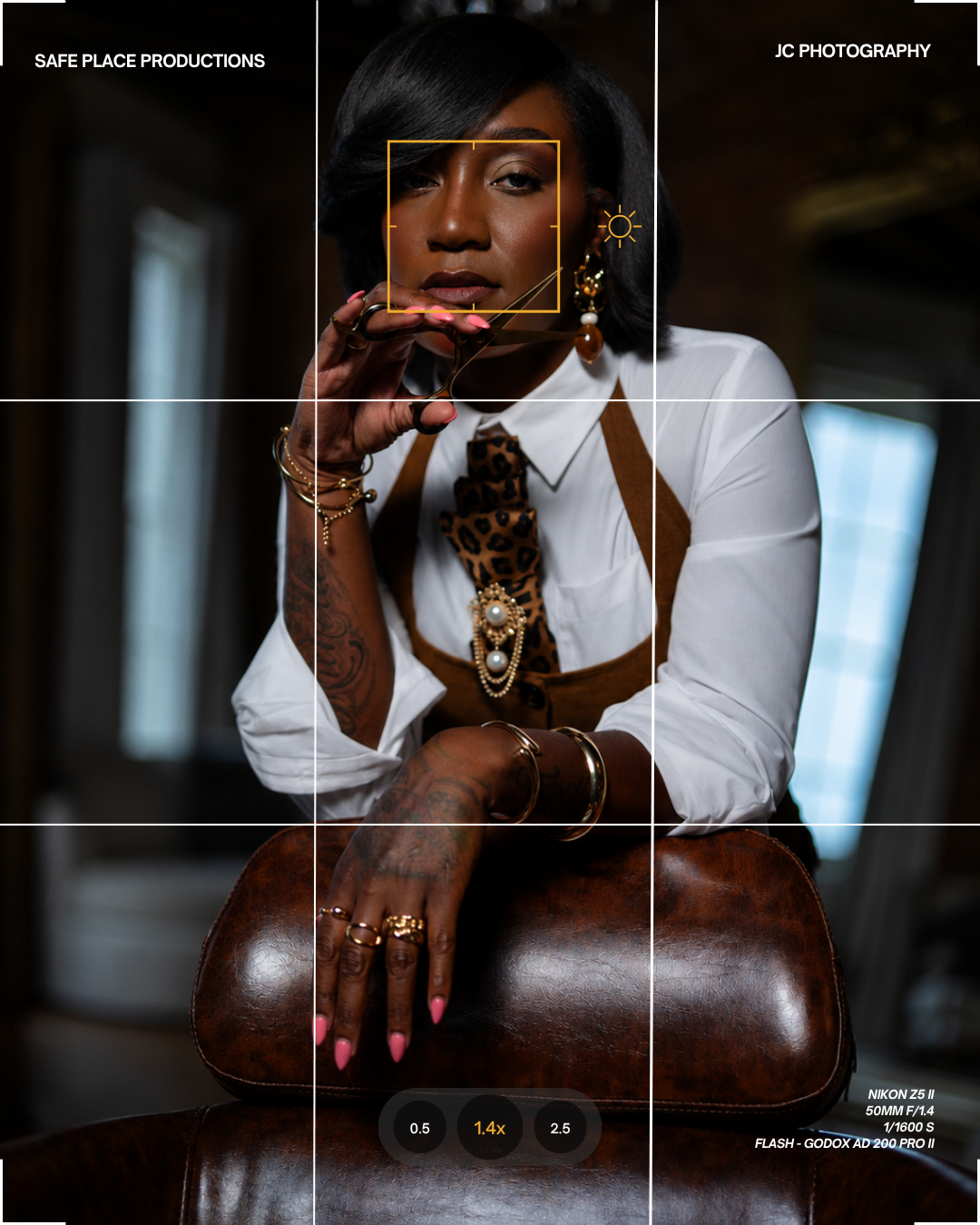 Portrait of a stylish Black woman with short hair, pink nails, tattoos, jewelry, and glasses, seated behind a leather chair in a photography studio