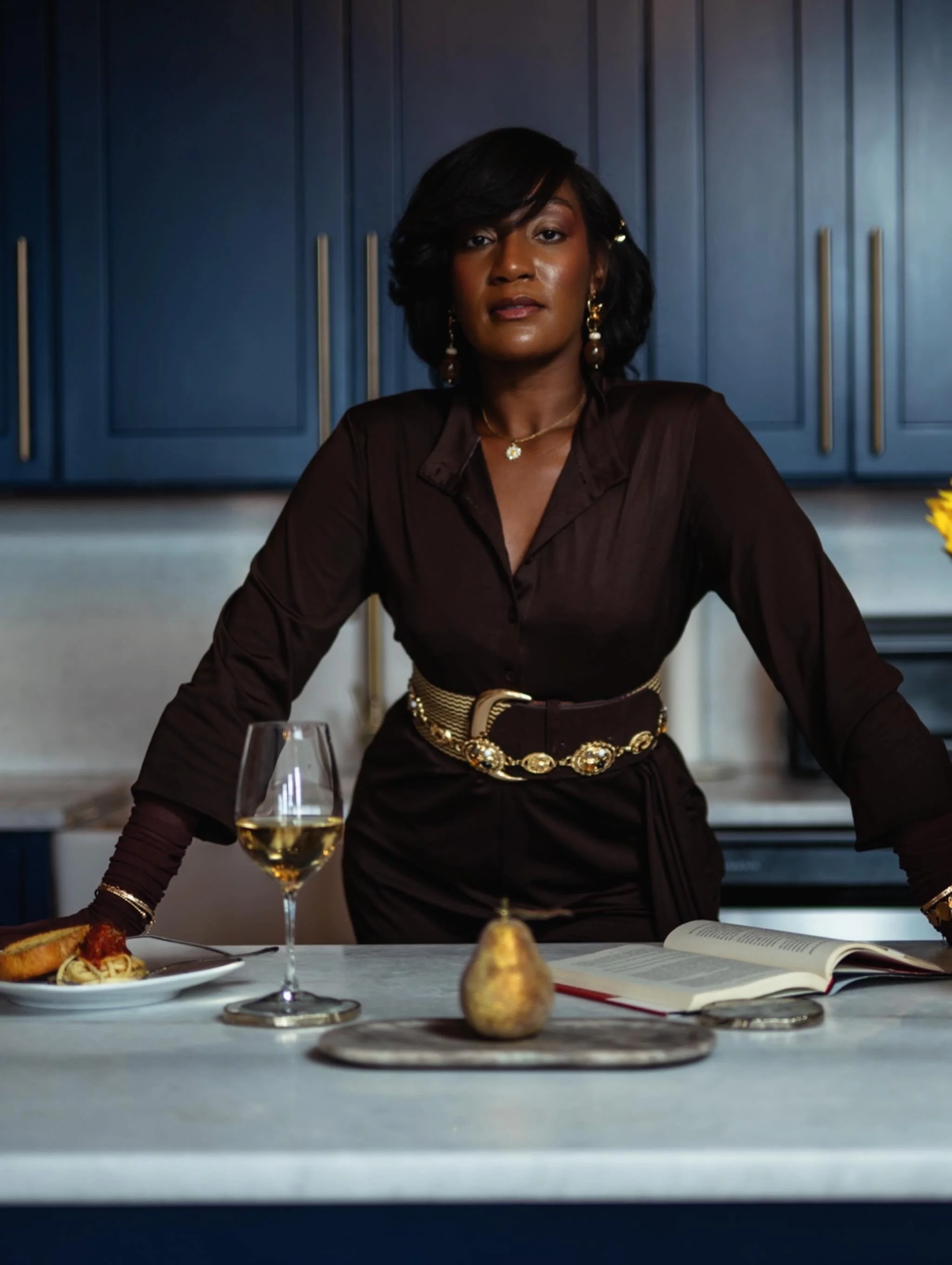 Black woman in an elegant dark outfit seated at a dining table with wine, food, and an open book