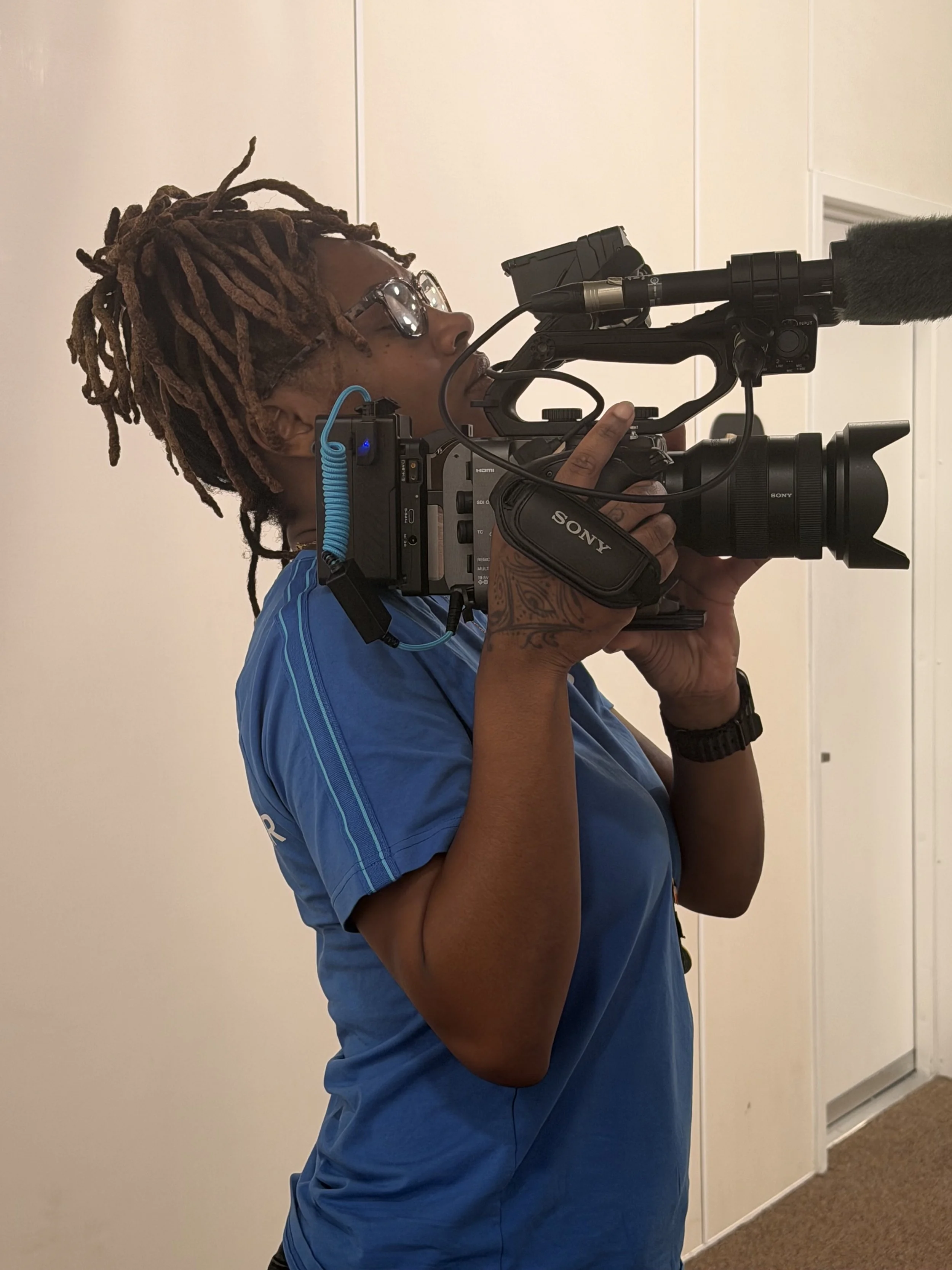 Person with dreadlocks and glasses operating a professional Sony video camera with a shotgun microphone.
