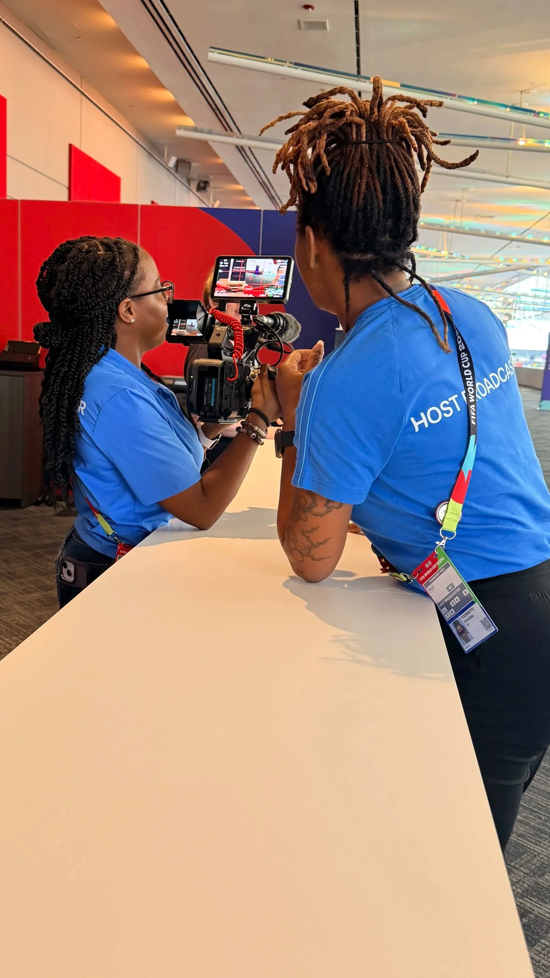 Two FIFA World Cup staff members in blue shirts film an interview inside a stadium media area.