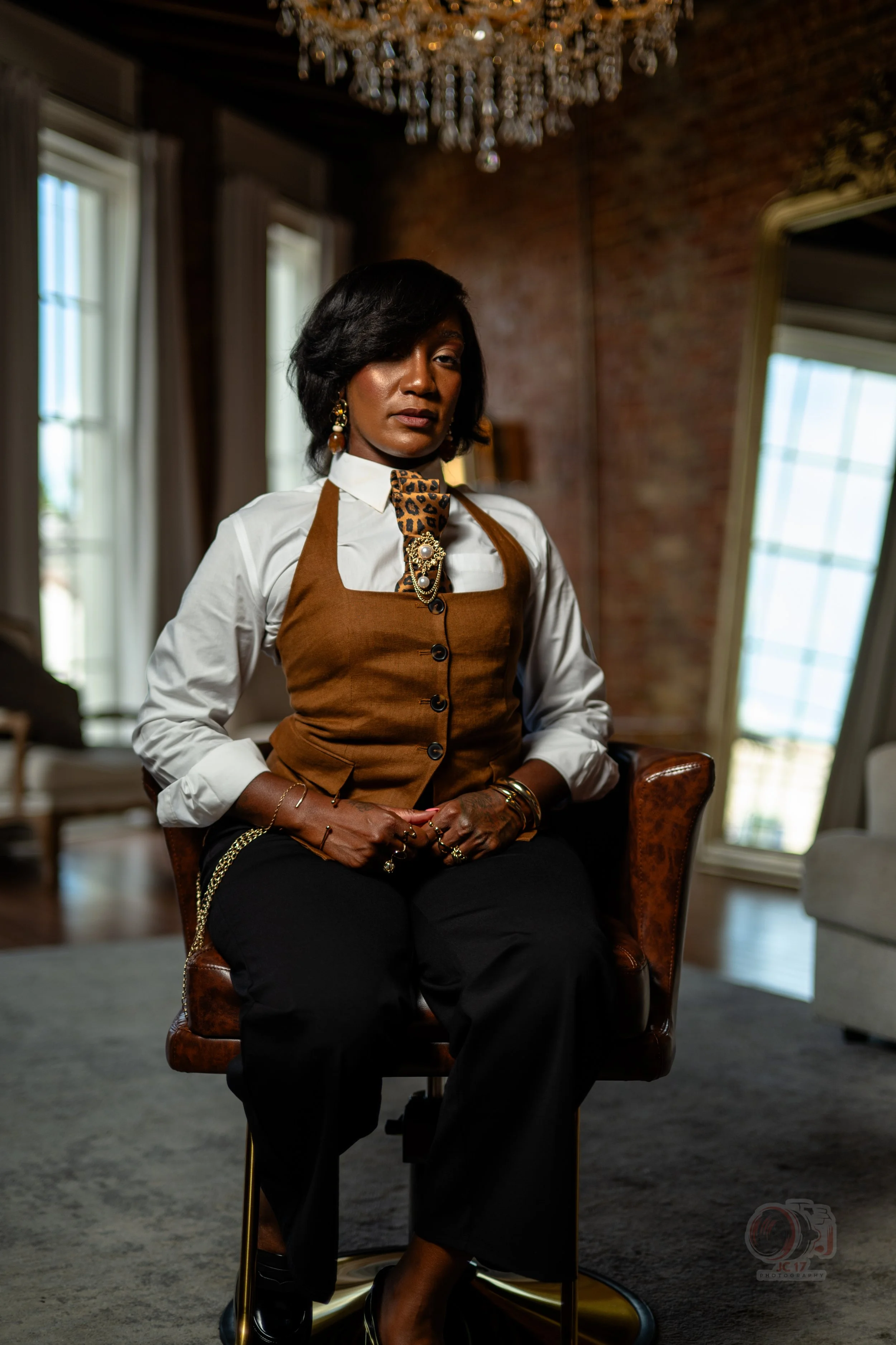 Black woman in a white blouse and brown vest seated in a leather armchair beneath a crystal chandelier
