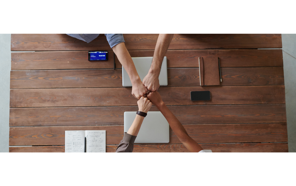Top view of four people clasping their hands together in a handshake over a wooden table with laptops, notebooks, and smartphones.