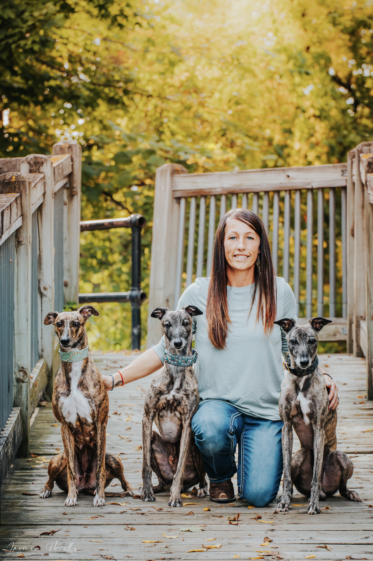 Woman kneeling with three brindle greyhound dogs on a wooden bridge surrounded by autumn foliage