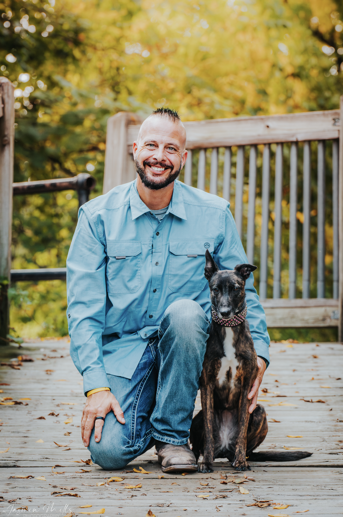 Smiling man in a blue shirt kneeling beside a black-and-brown dog on a wooden deck in autumn