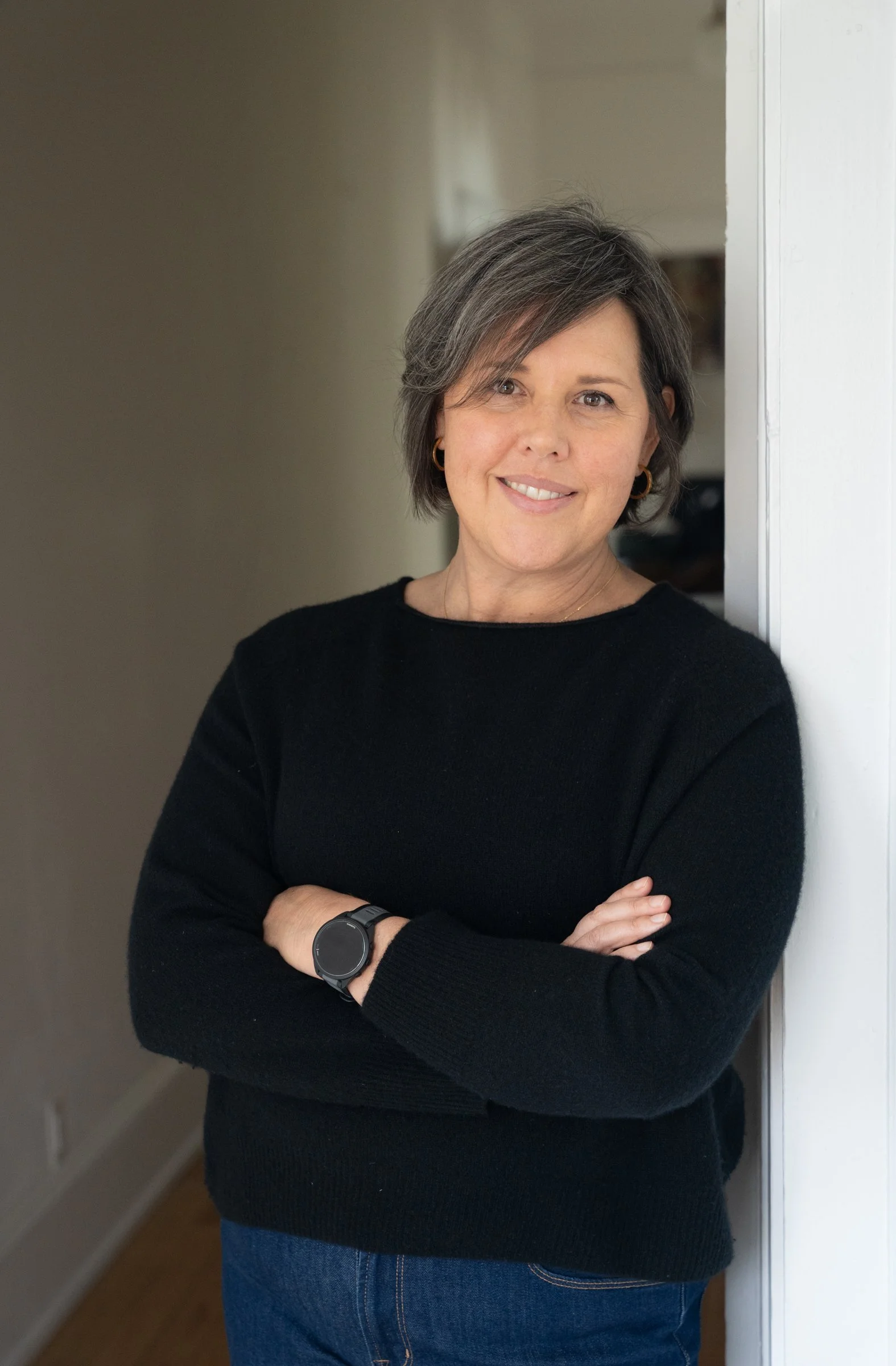 Smiling middle-aged woman in a black sweater and jeans standing with crossed arms in a doorway.