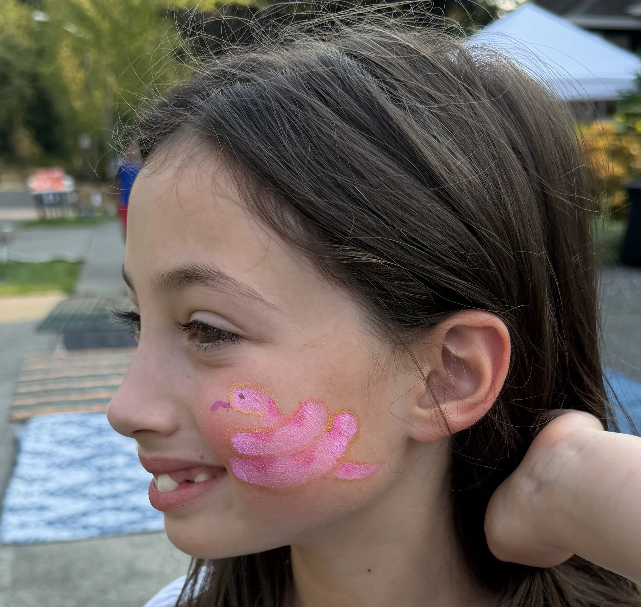 Close-up of a smiling girl with dark hair and a colorful pink-and-orange flamingo face painting outdoors