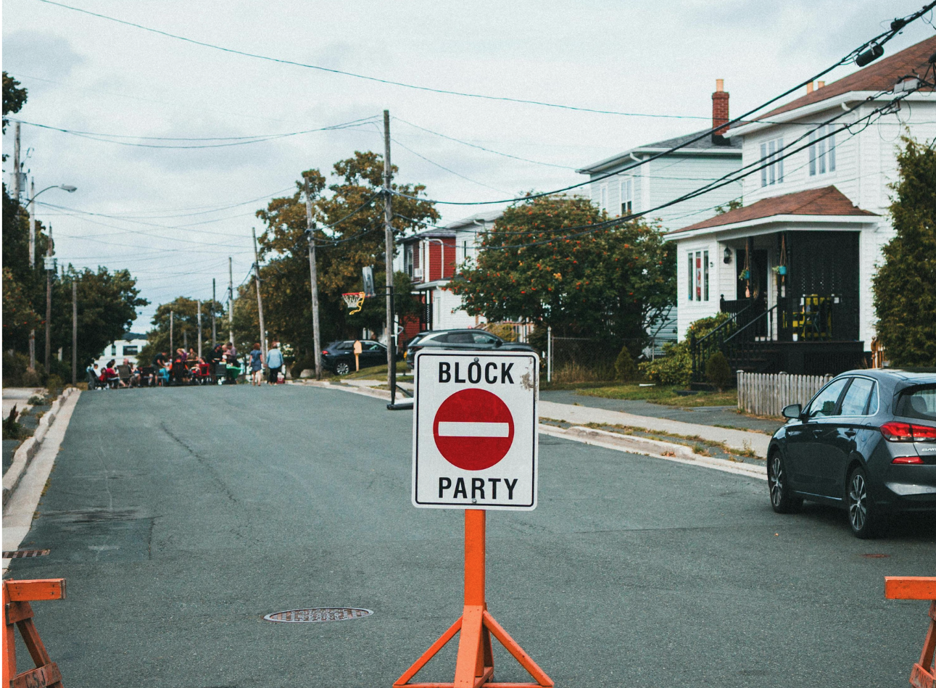 Residential street closed for a block party with a temporary “BLOCK PARTY” road sign, houses, parked car, and people gathering in the distance.