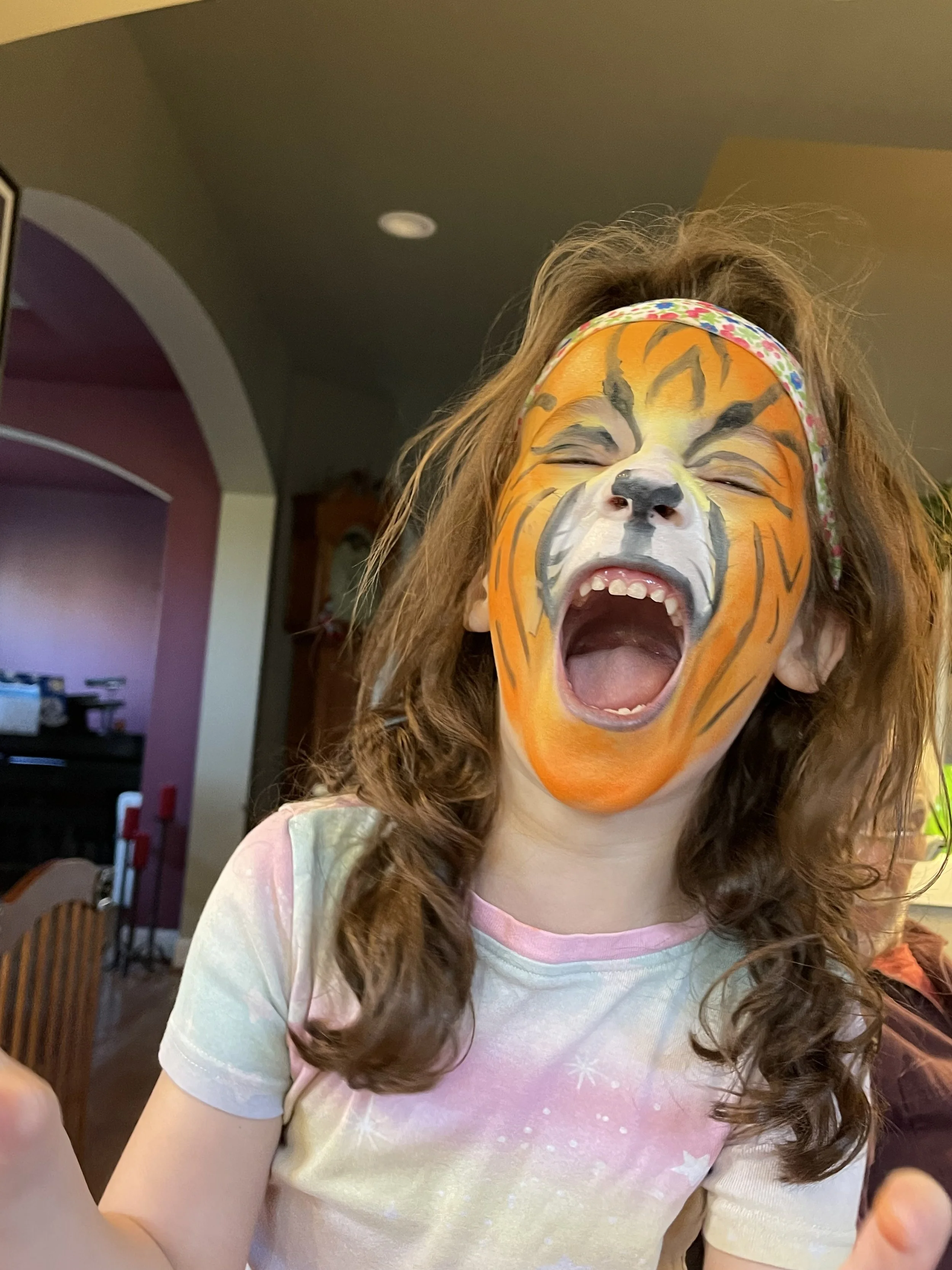 Child with orange tiger face paint, a floral headband, and long brown hair smiling at the camera.